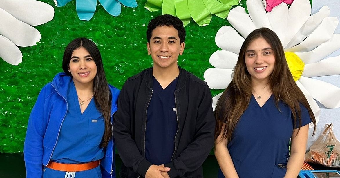 Three smiling healthcare workers stand together, blue scrubs, flower backdrop.