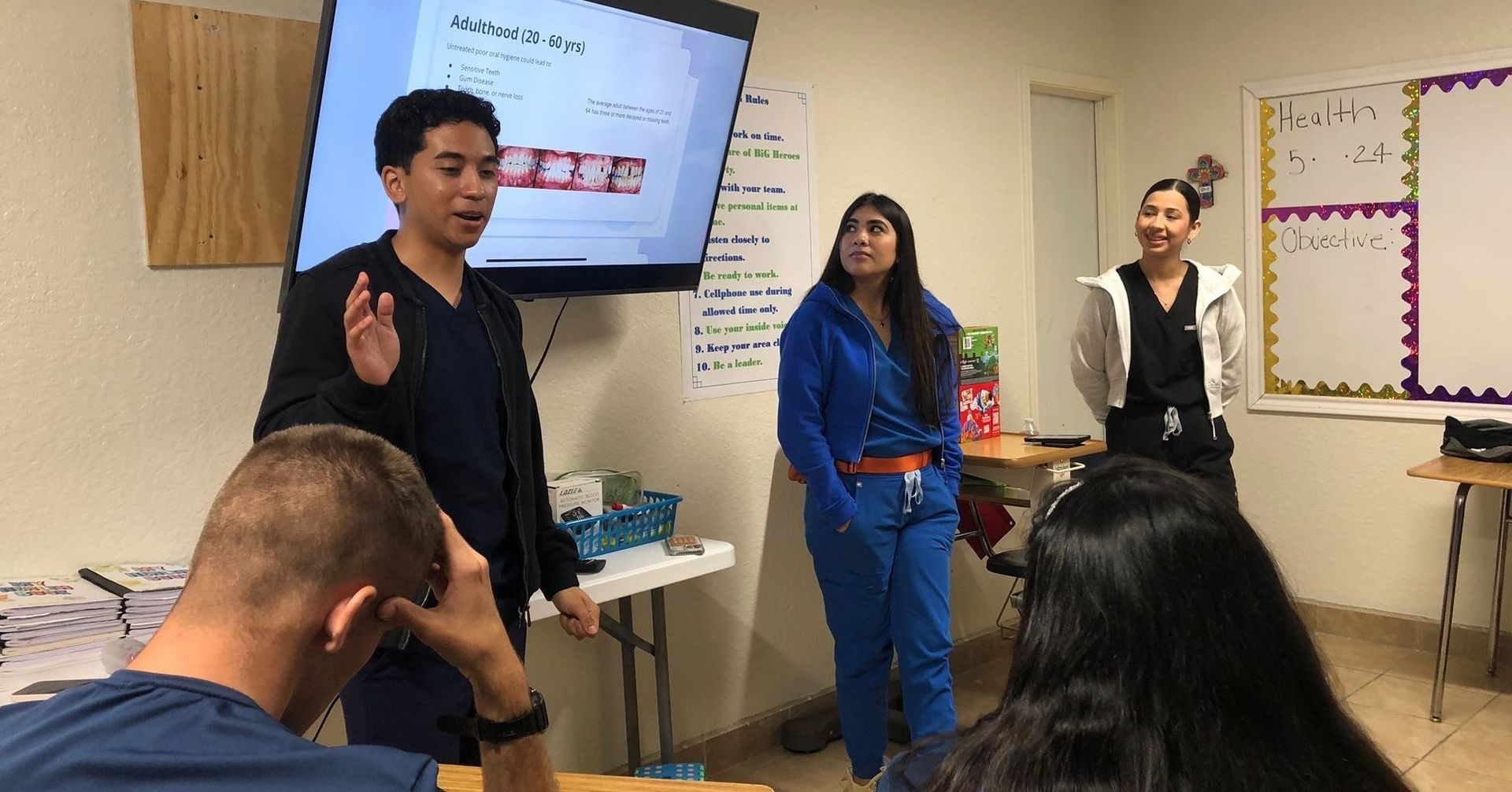 A young man presents in a classroom. Two others stand nearby. Students sit at desks.