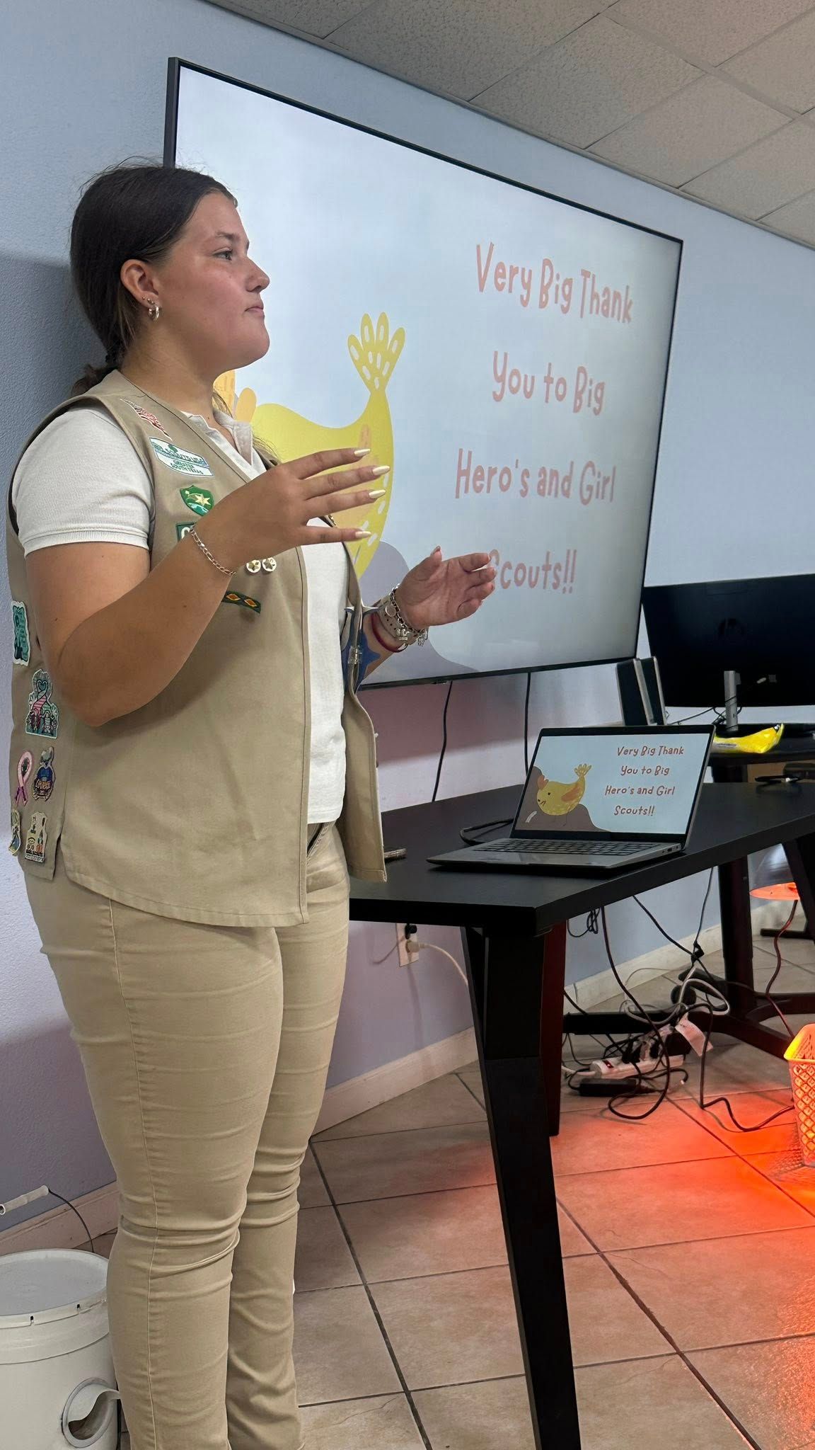 Woman giving a presentation, beige attire, in front of a screen with text. Interior setting.