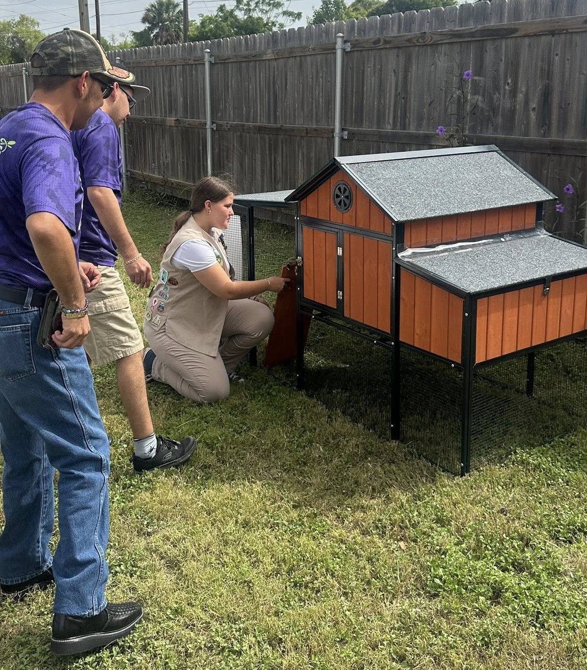 Three people assemble a brown and black chicken coop in a backyard.