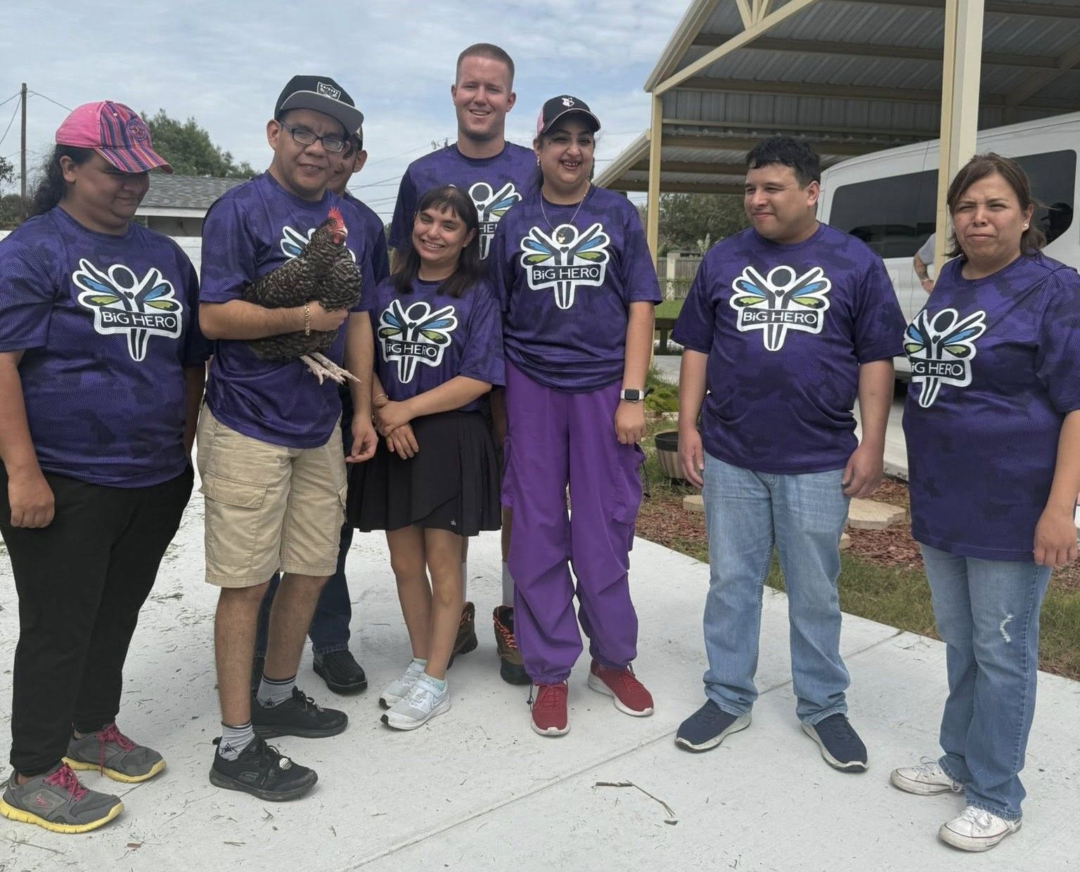Group of people in purple shirts with logo, pose outside with a chicken.
