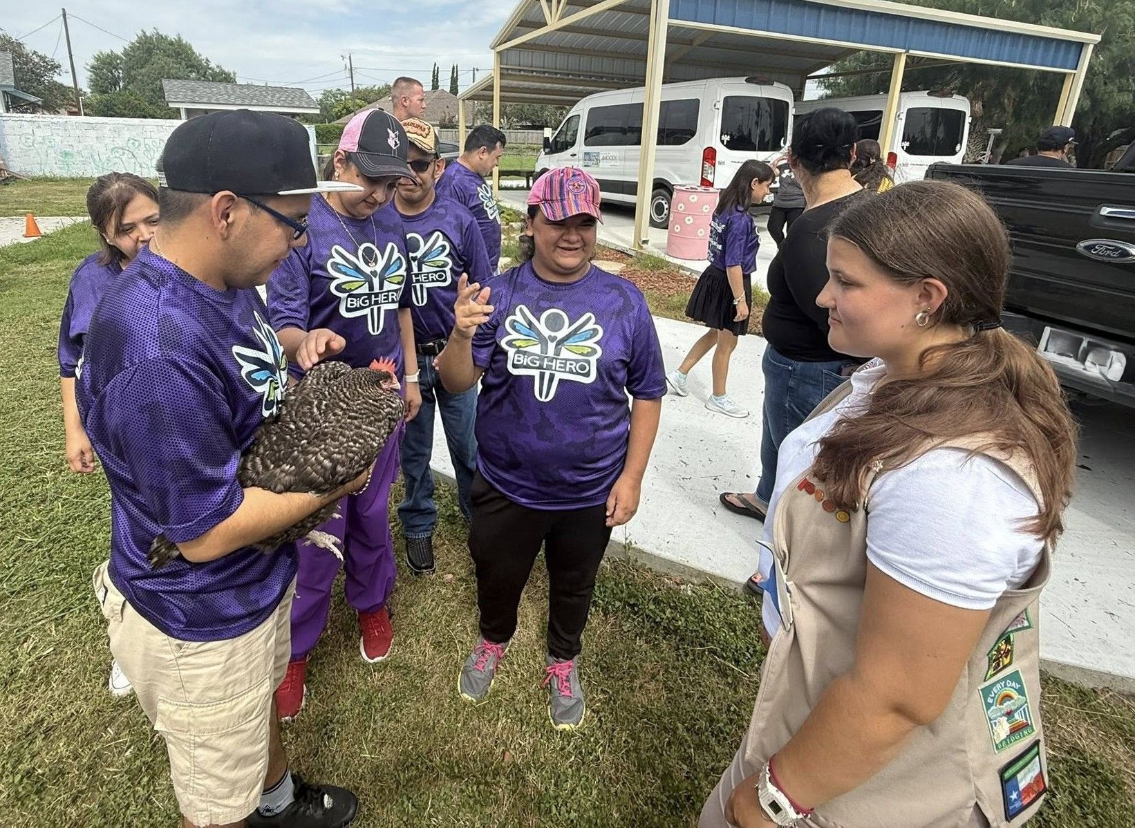 A group of people, some in purple shirts, gather outdoors to see a person holding a chicken.