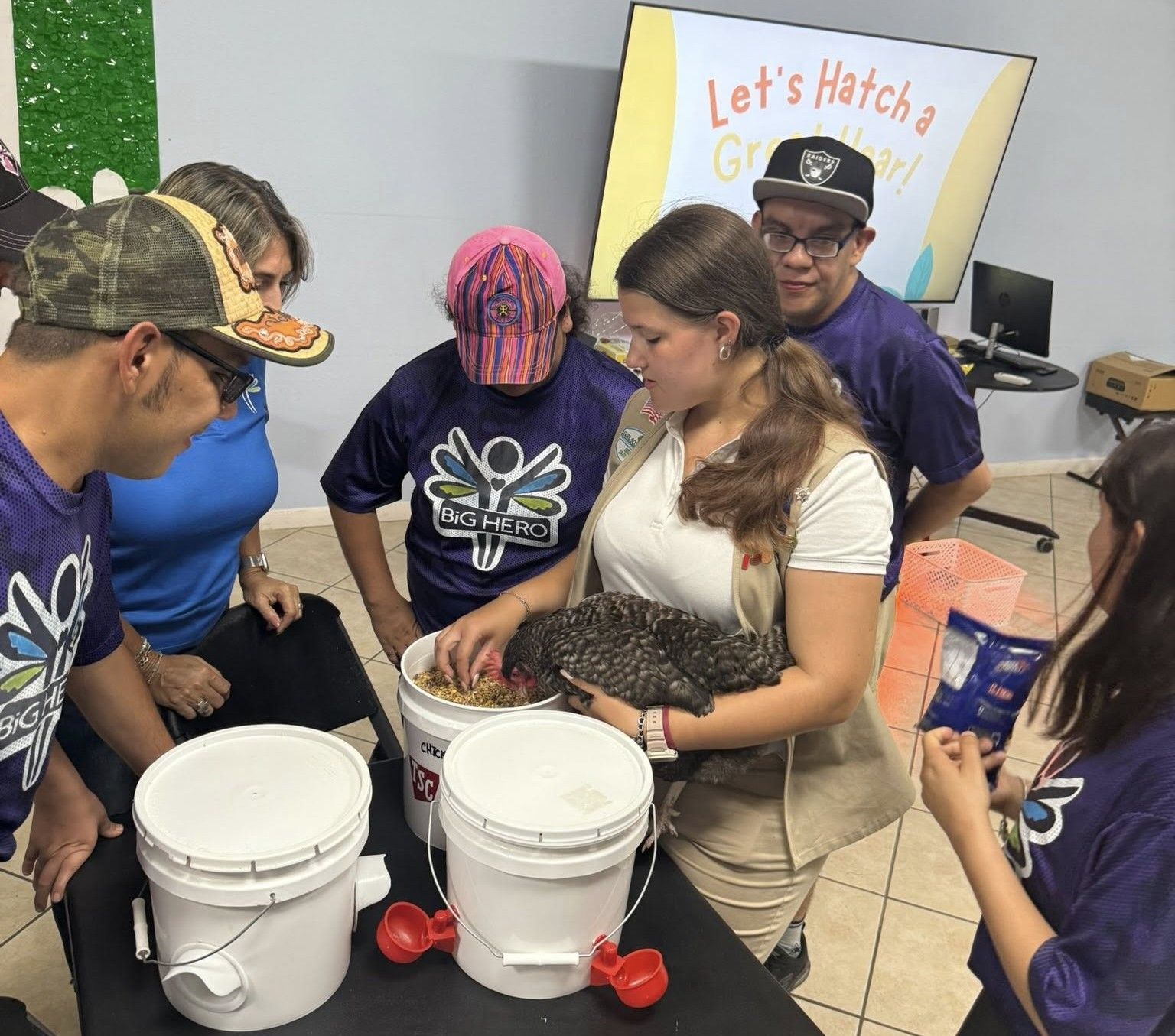 Group of people observing a woman holding a chicken, looking at chicken feed in buckets.