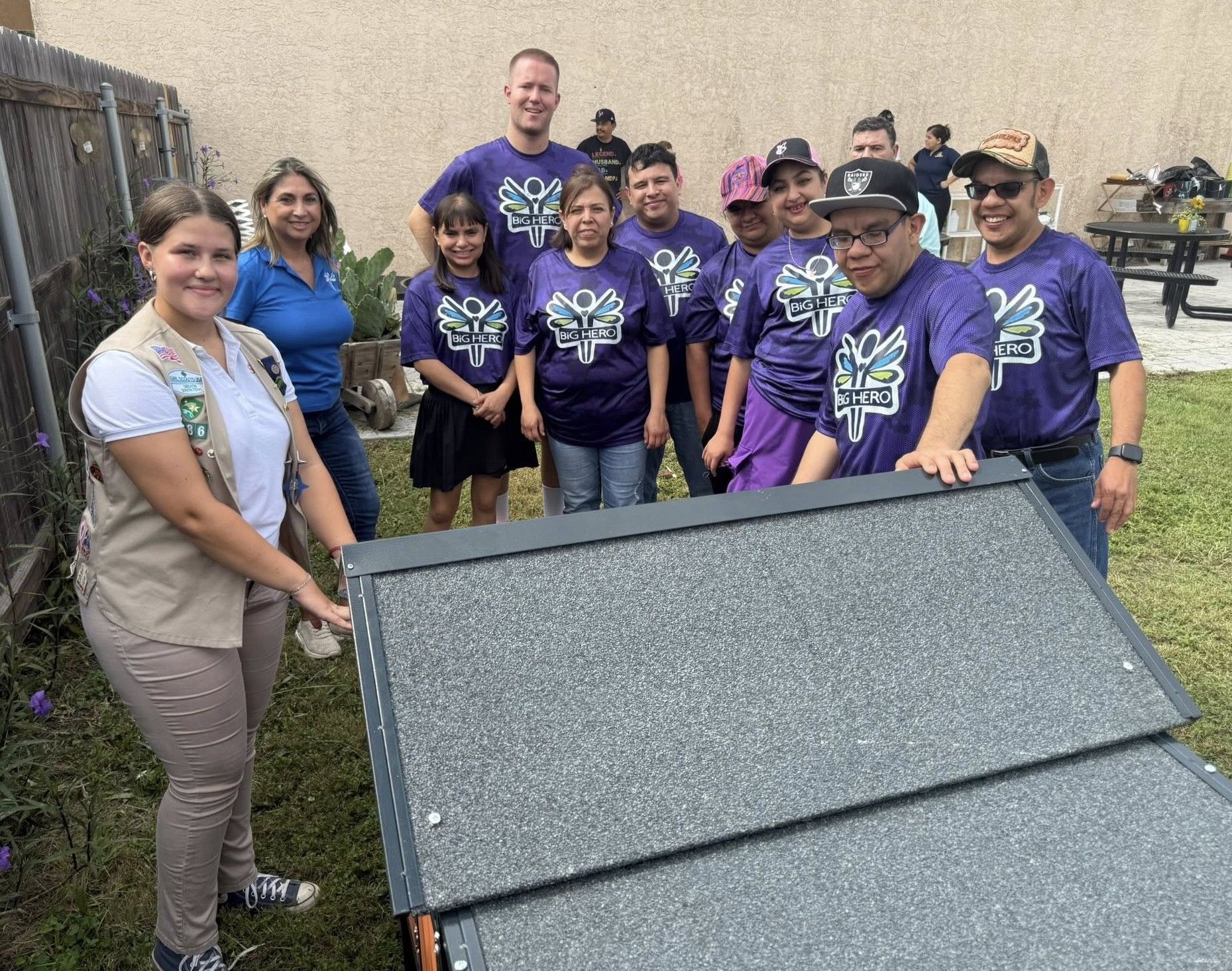 Group of people assembling a structure outdoors. Some wear purple shirts with a tree design, smiling.