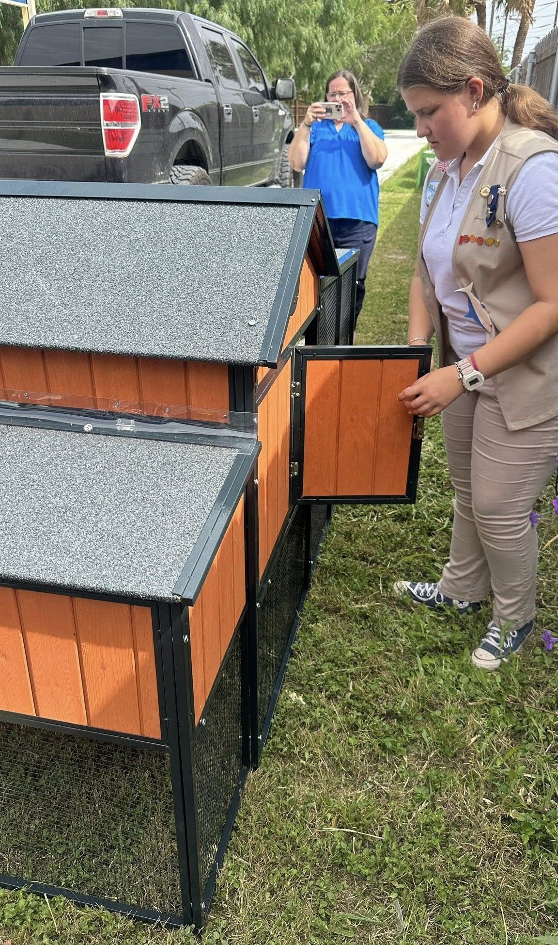 Girl Scout opens the door of a wooden chicken coop in a yard.