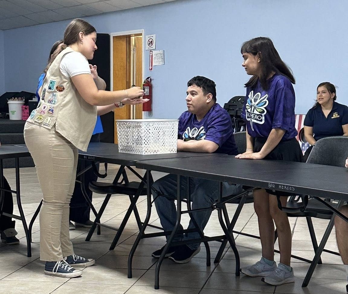 Girl Scout hands items to two people at a table; inside, casual setting.