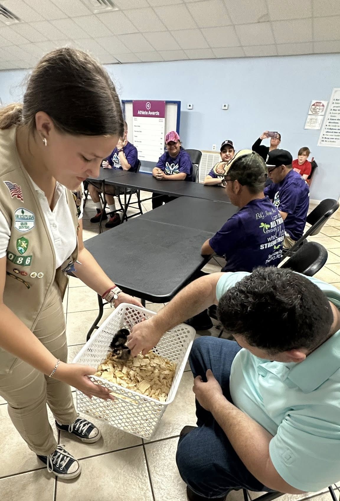 Girl Scout serves snacks to people seated at a table in a room, some in purple shirts.