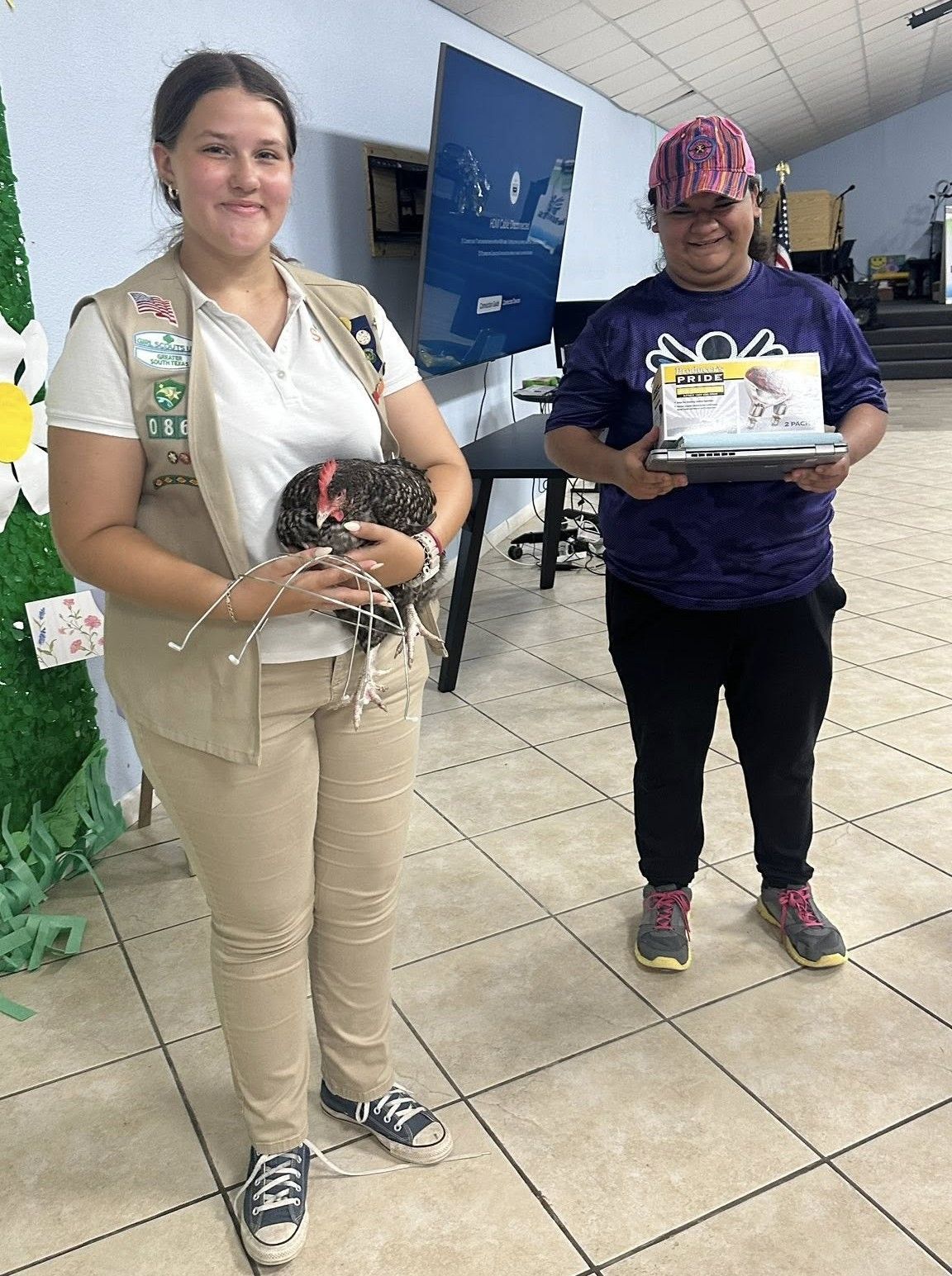 Girl Scout holding a chicken, smiles at the camera; another person holds a framed award. Indoor setting.