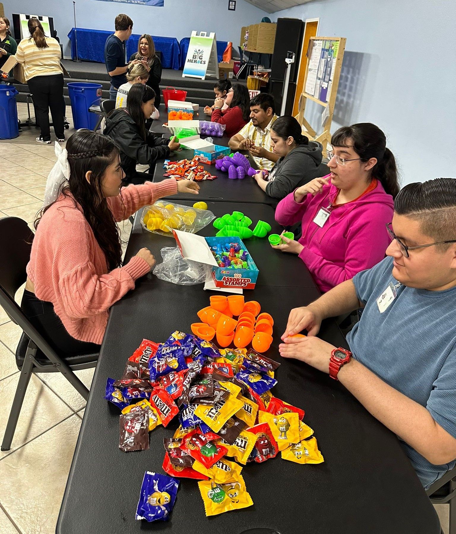 People at a table sorting small toys into Easter eggs; inside an event space.