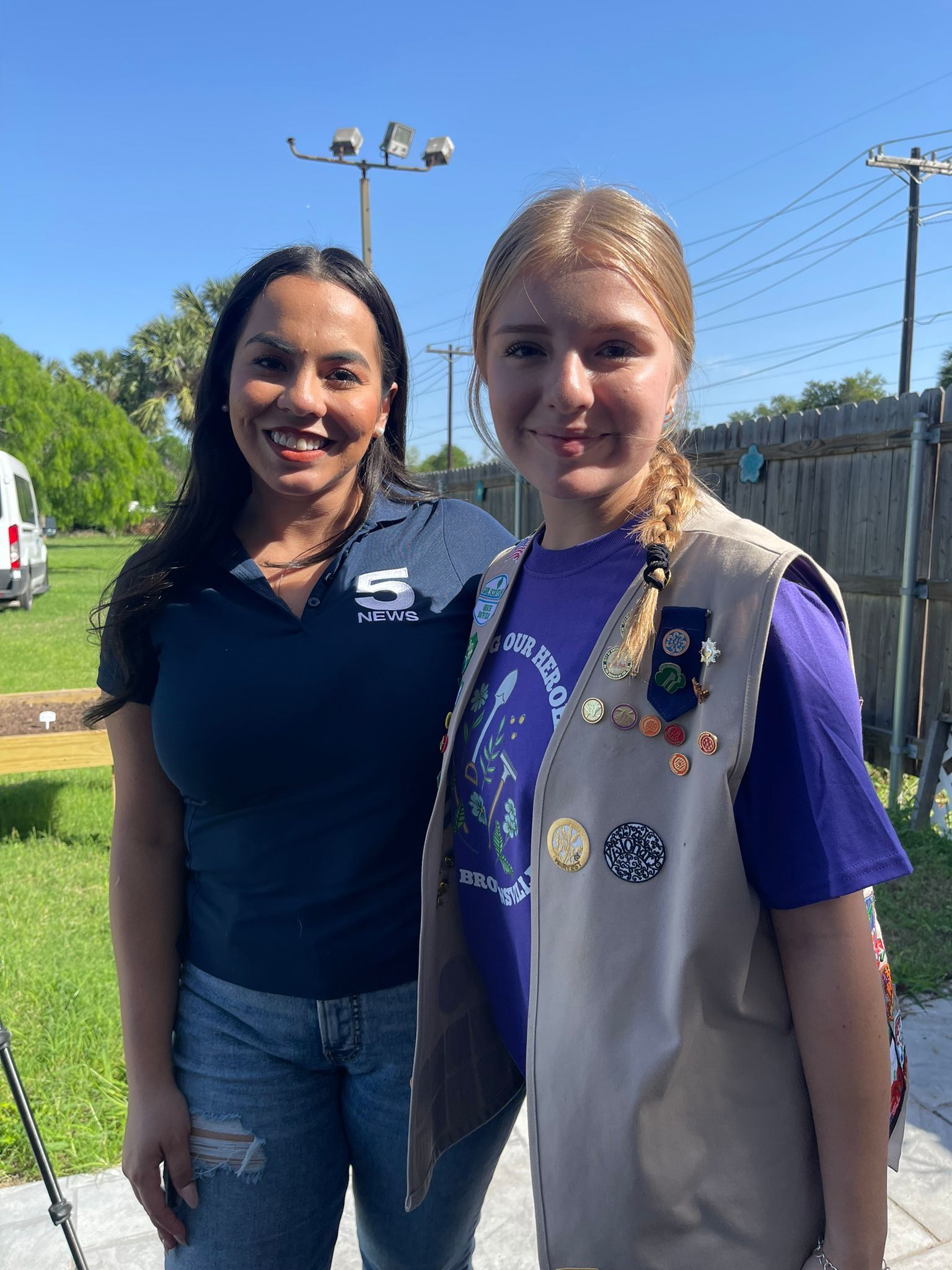 Woman and Girl Scout smiling outdoors. Woman in blue shirt, girl in vest, sunlight.