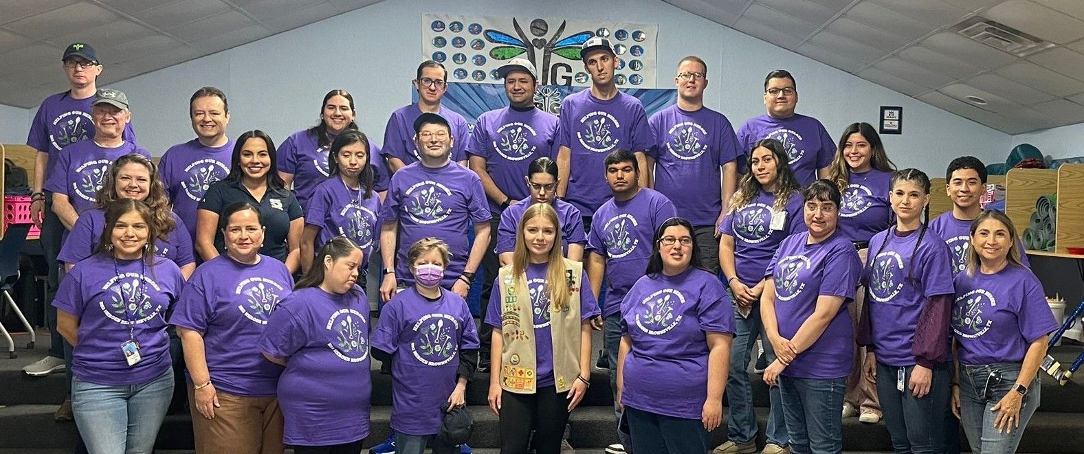 Group of people in purple t-shirts posing indoors, with a mural in the background.