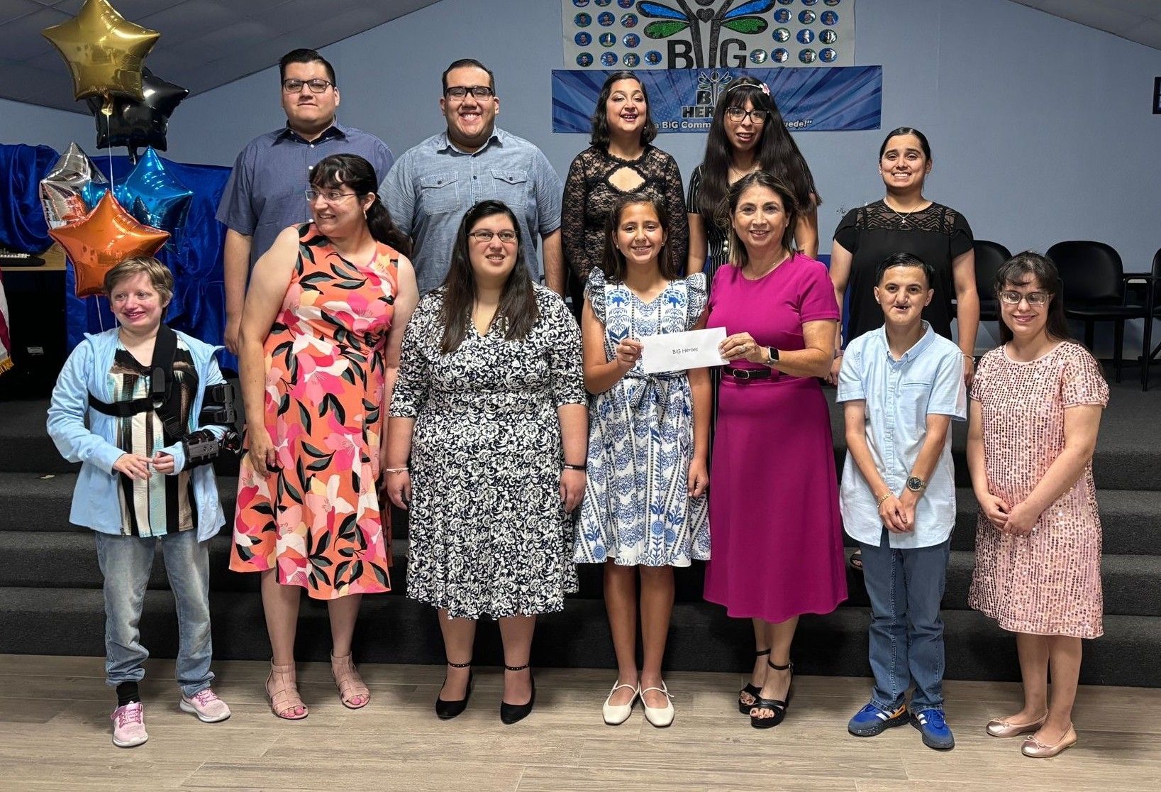 Group of people, some holding a check, posing on stairs in a brightly lit room, for an event.