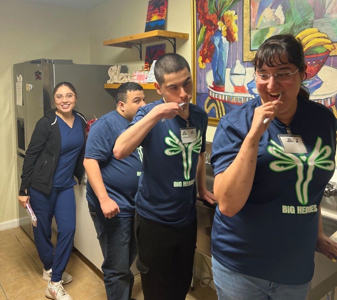 Four people brushing teeth in a kitchen. Woman in scrubs smiles, men and another woman brush teeth, all in matching shirts.