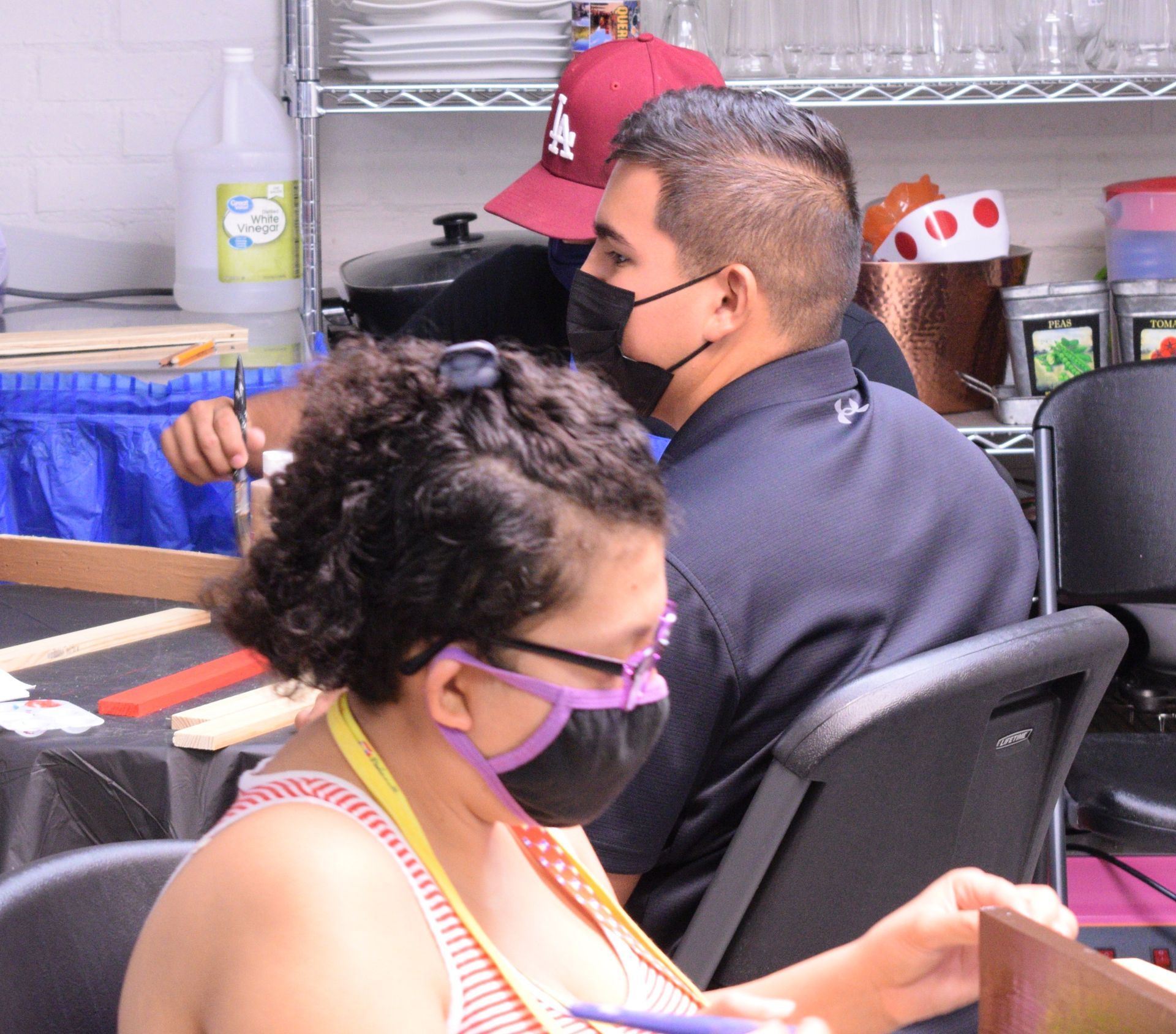People wearing masks in a workshop setting. One person paints, others observe.