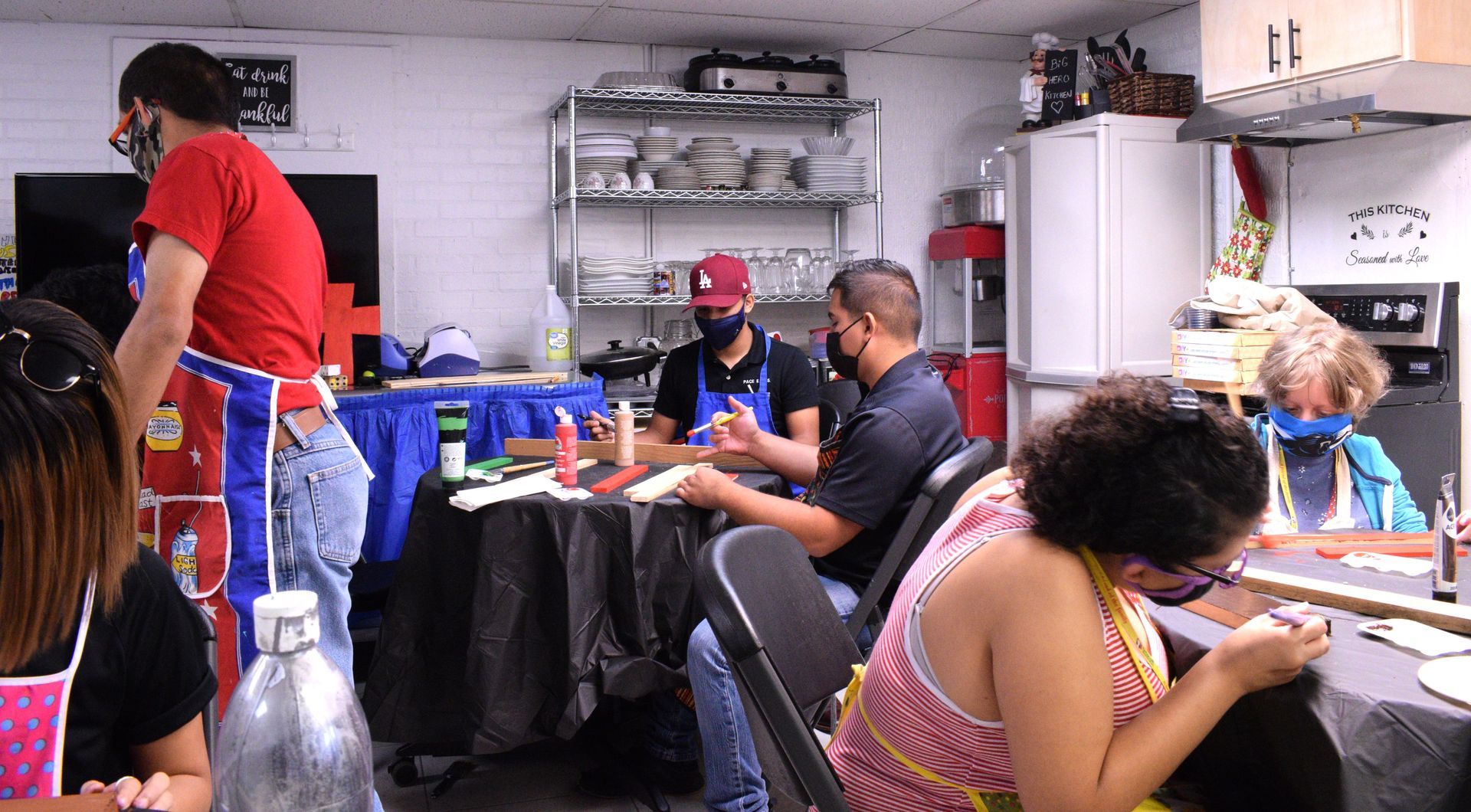 People crafting at tables in a room; a man in a red shirt assists.