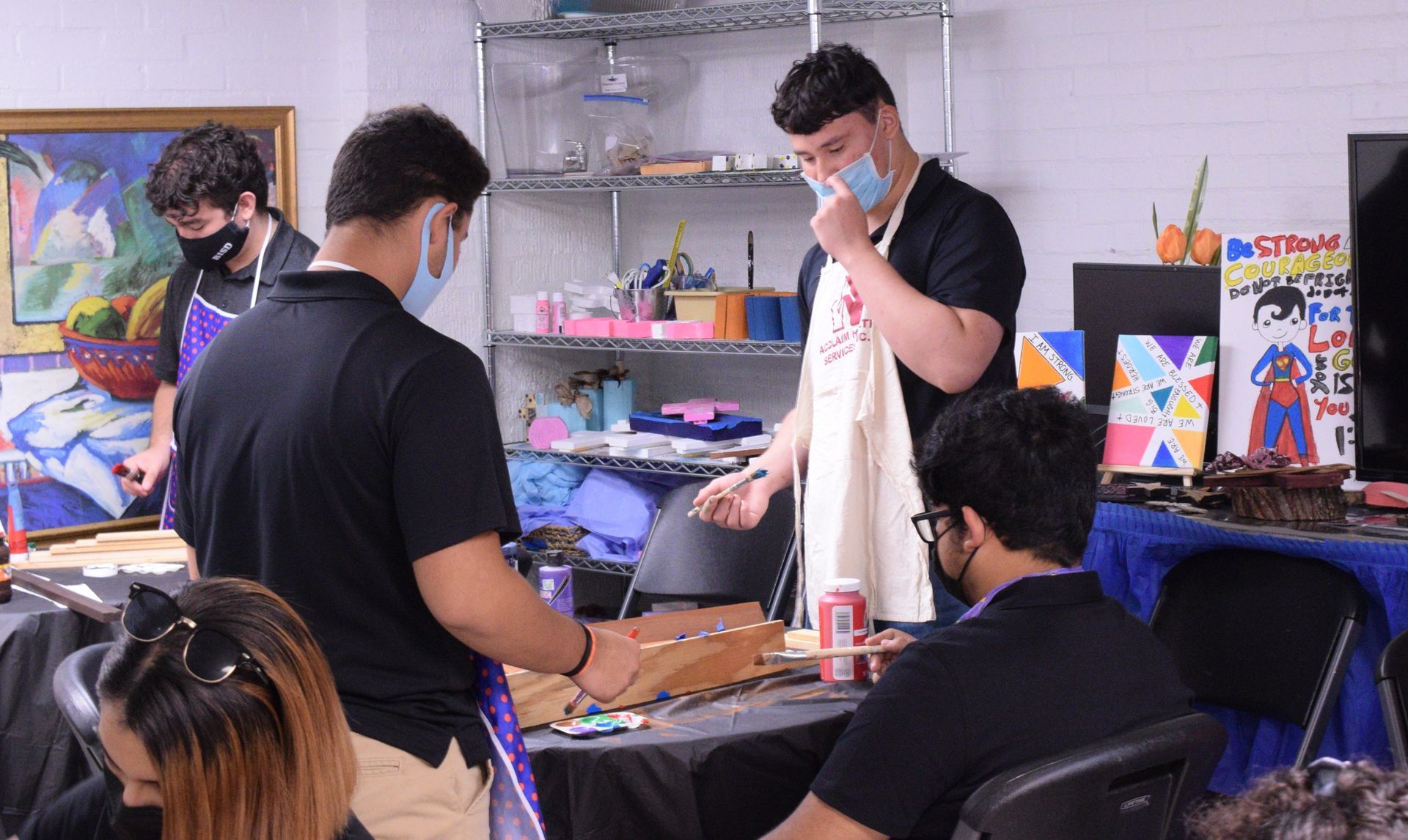 Four people painting at a table, wearing masks and aprons. Art supplies and a colorful painting in the background.