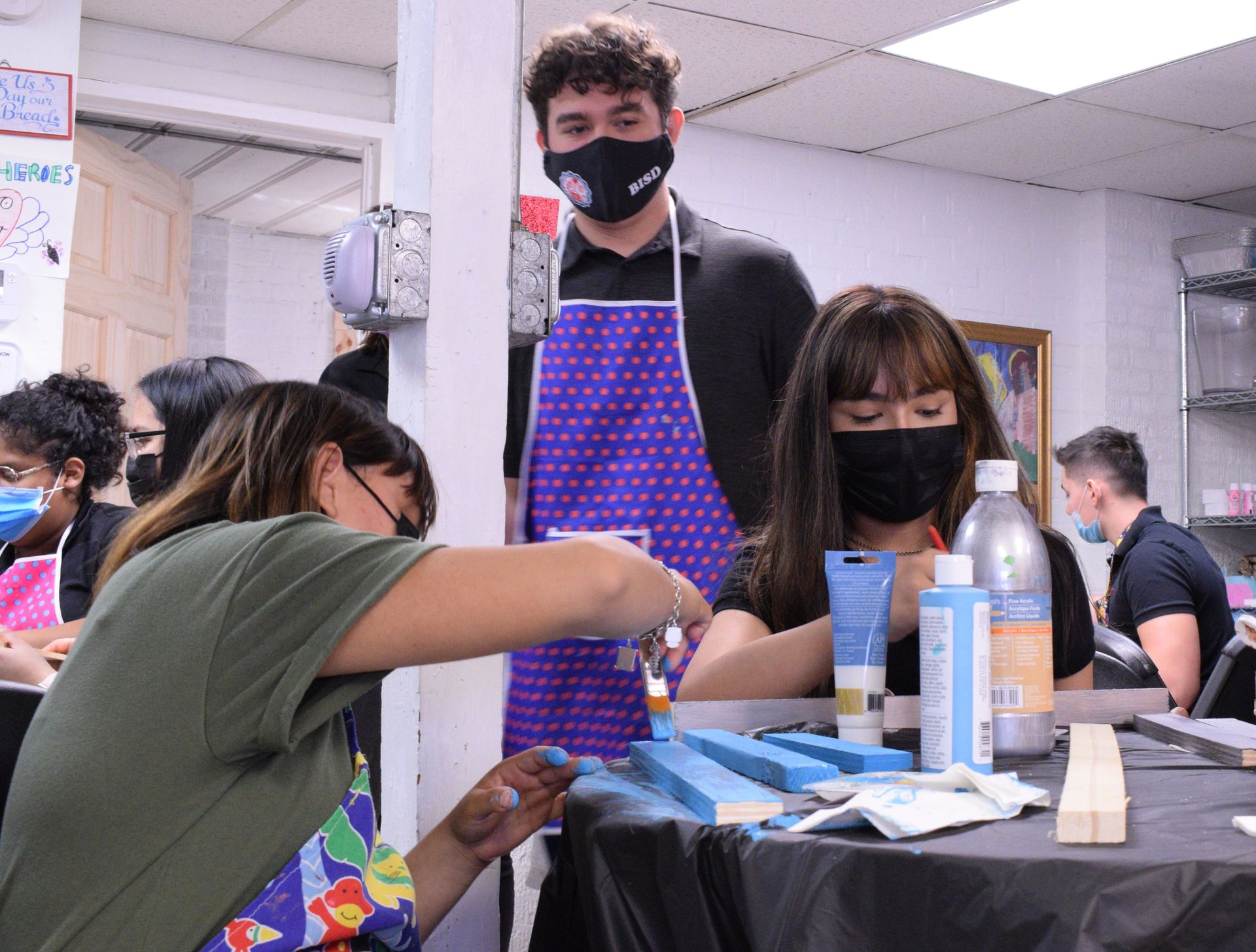 People painting wooden planks at a workshop; instructor watches. Everyone wears masks and aprons.