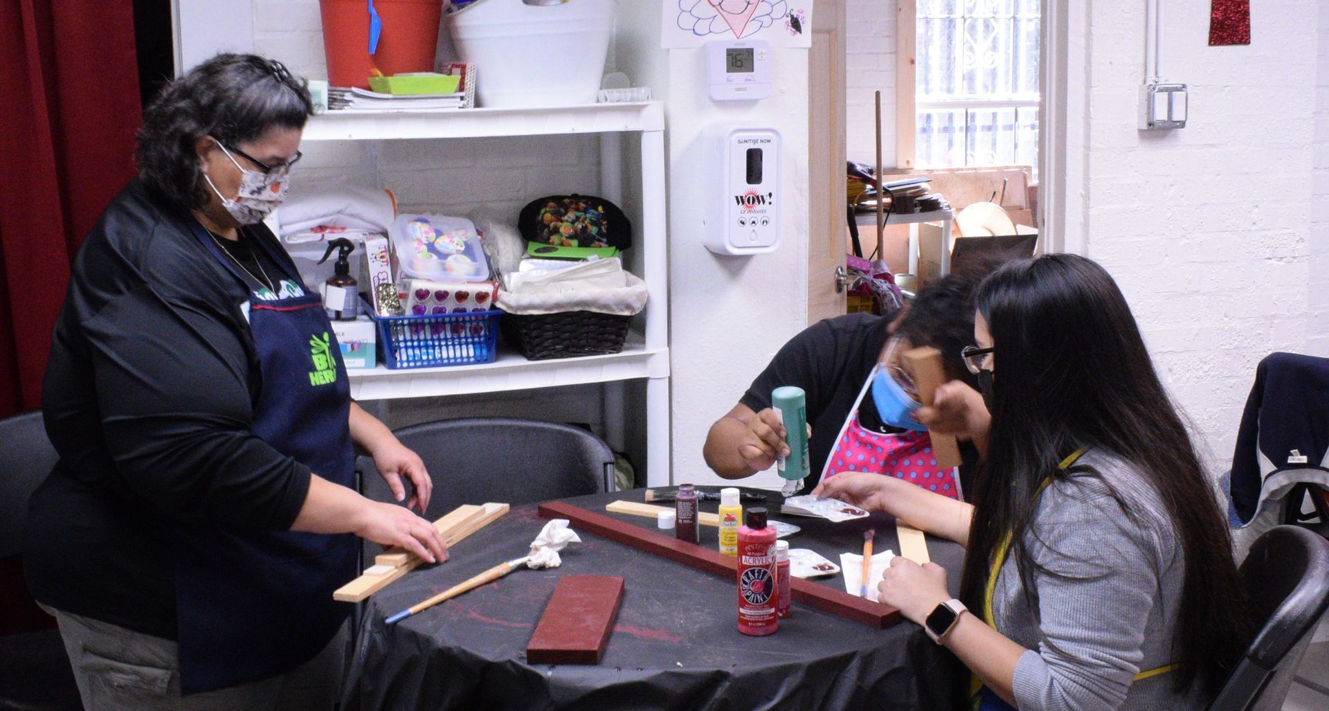 Woman in apron assisting two others painting wood pieces at a table. Indoors.