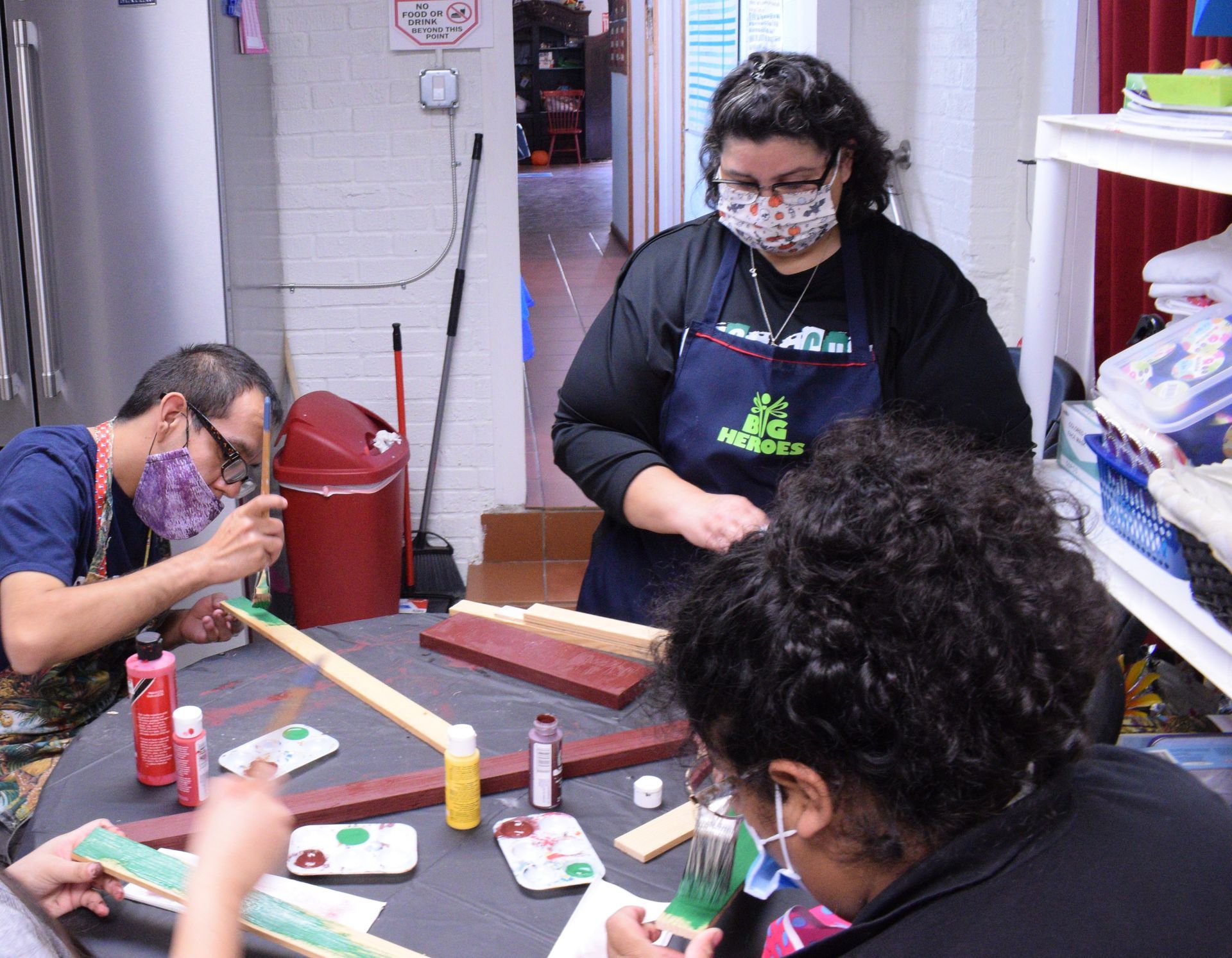 People painting wooden objects at a table. One woman oversees, wearing an apron and mask.