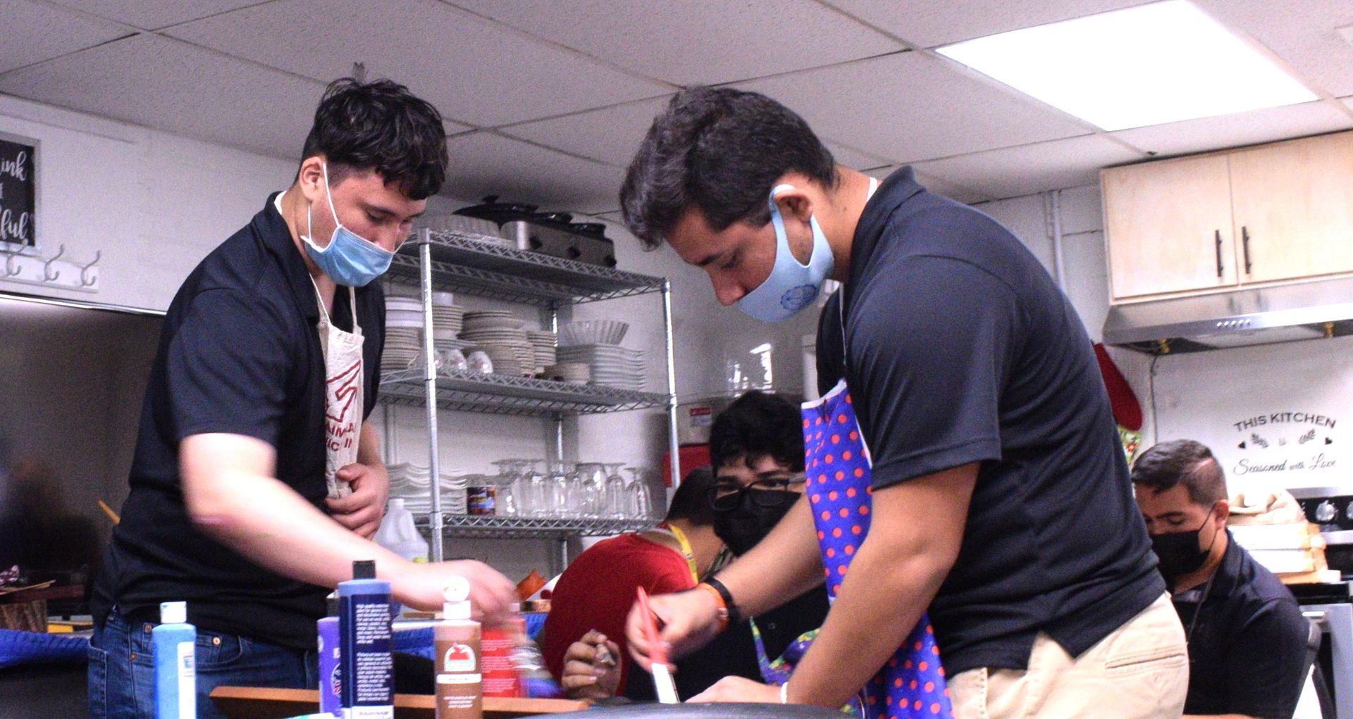 People wearing masks cooking in a kitchen. One stirs a pot, others look on.