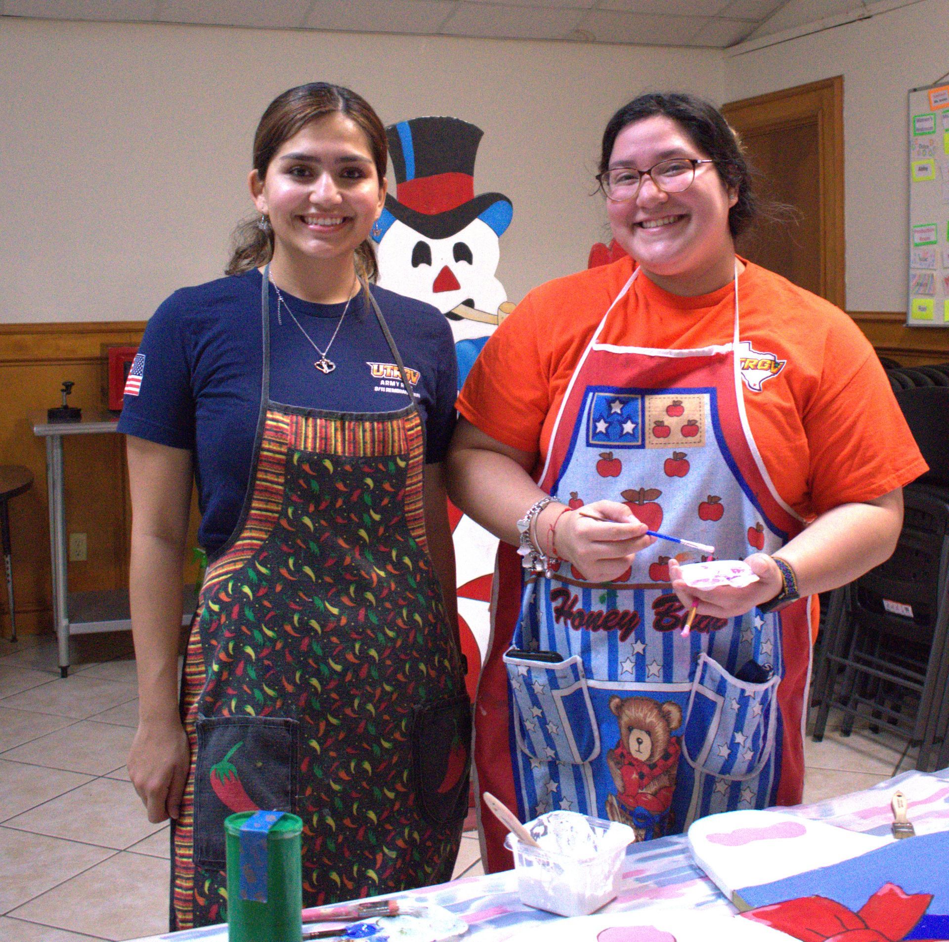 Two women wearing aprons smiling while painting in a room. A snowman stands in the background.