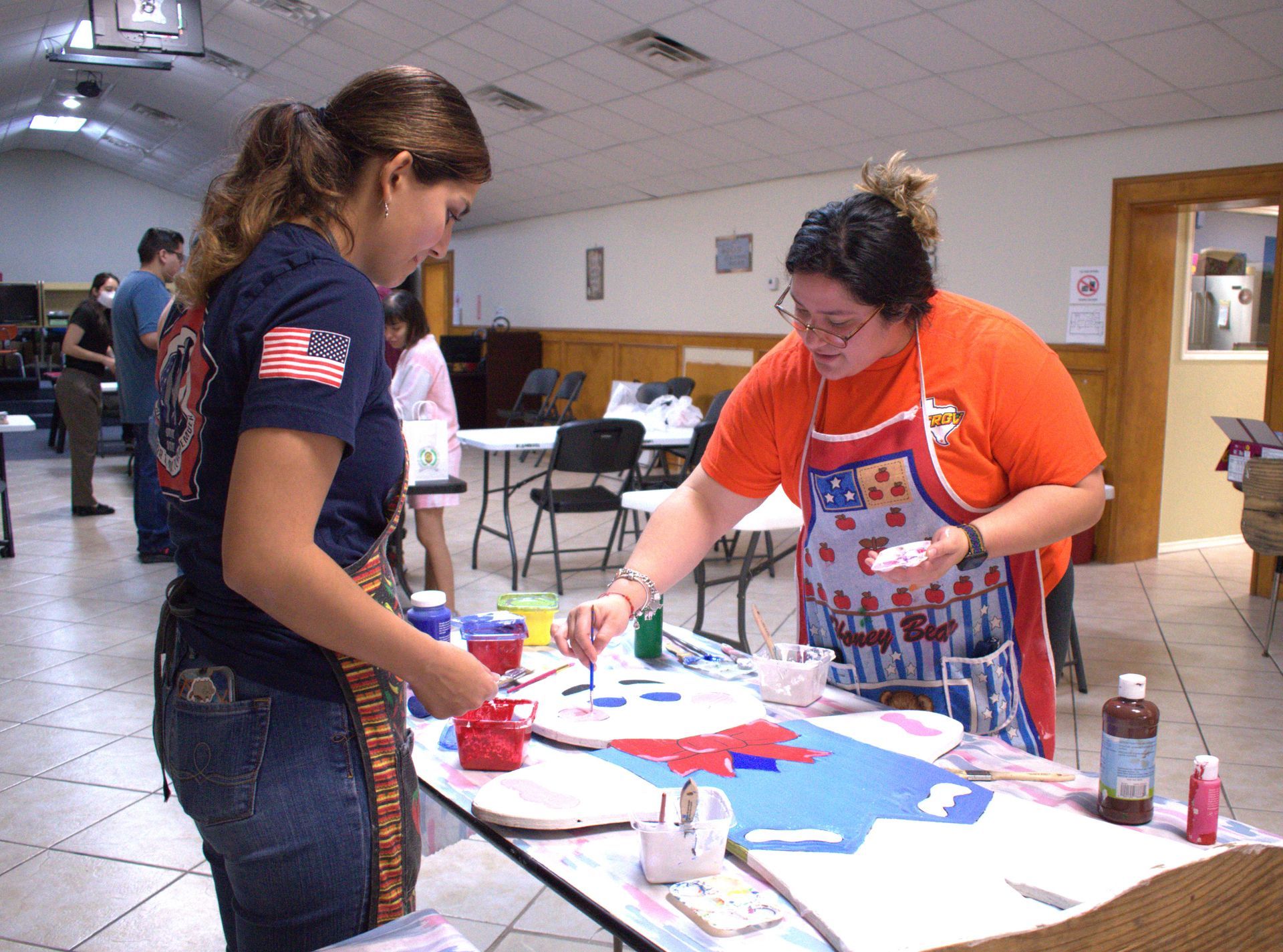 Two women painting a craft project at a table in a room. One wears a red, white, and blue apron.