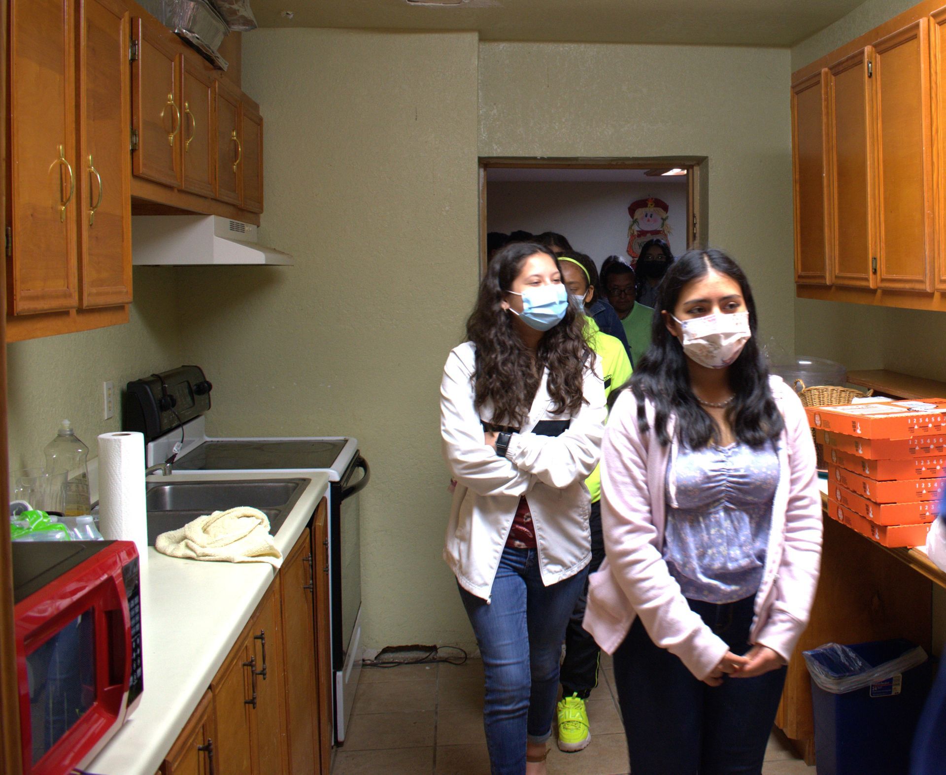 People wearing masks walk through a kitchen with wooden cabinets.