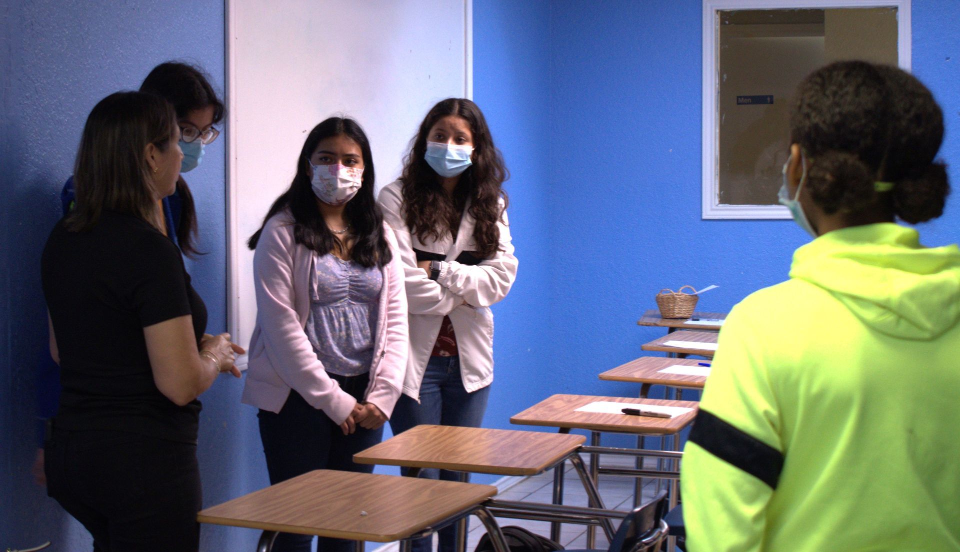 Students and teacher in a classroom, wearing masks. Blue walls, desks, whiteboard.