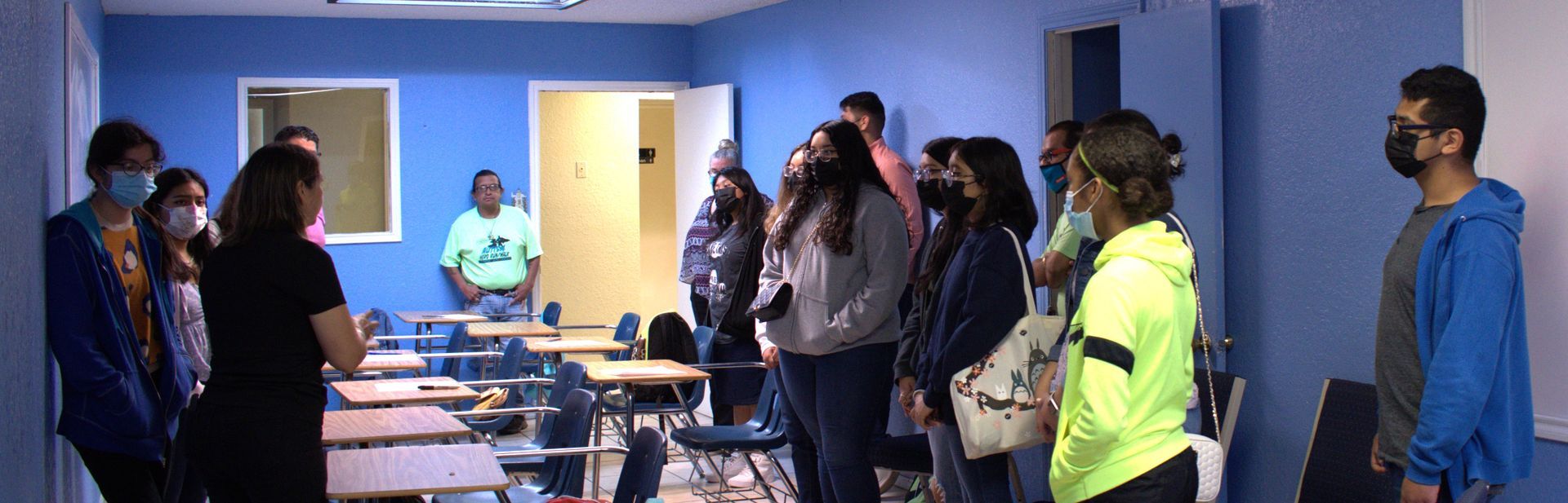 A group of students in a classroom, wearing masks. Some are looking upwards and the room is blue.