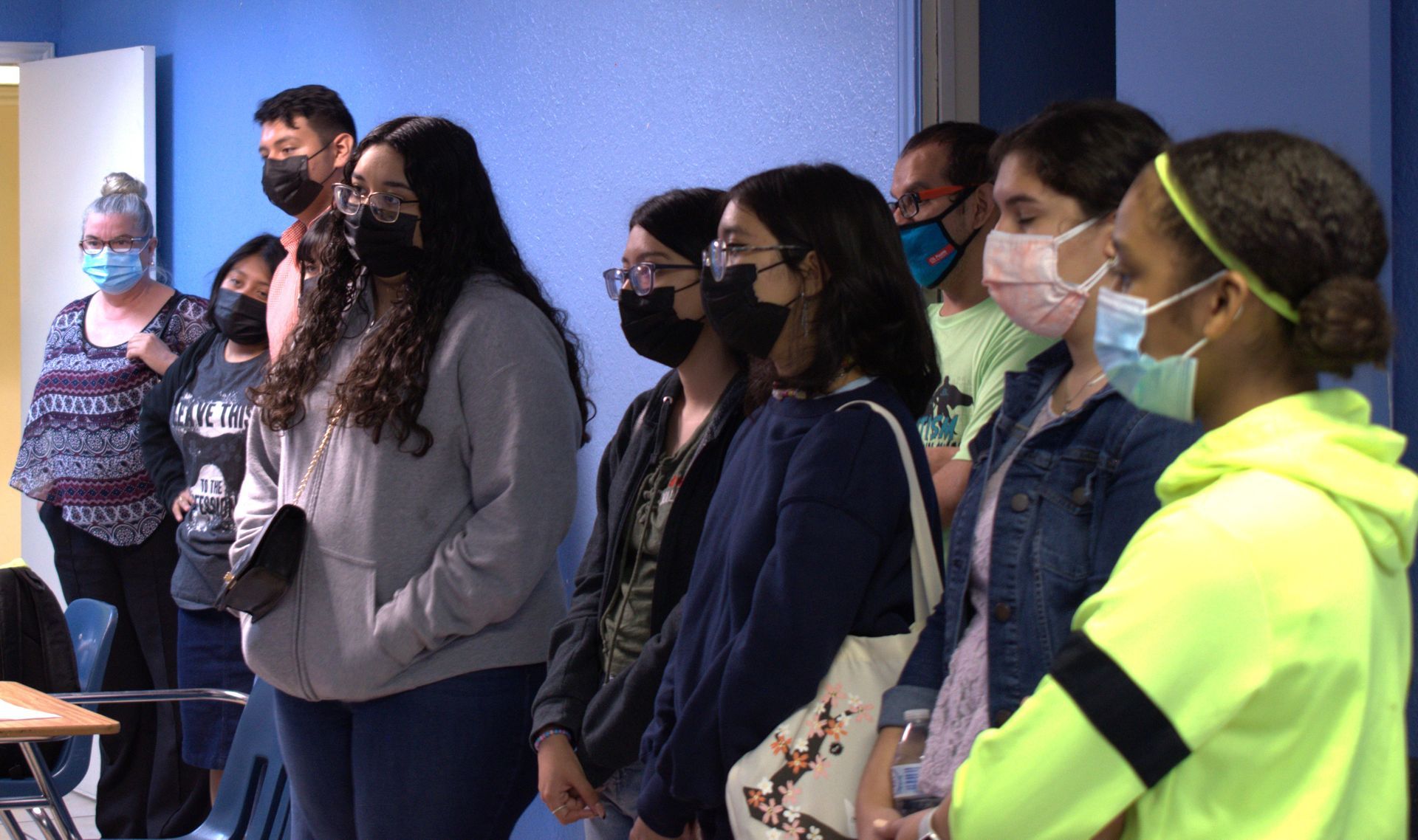 Group of people wearing masks stand in a classroom; some look forward, one woman has light blue hair.