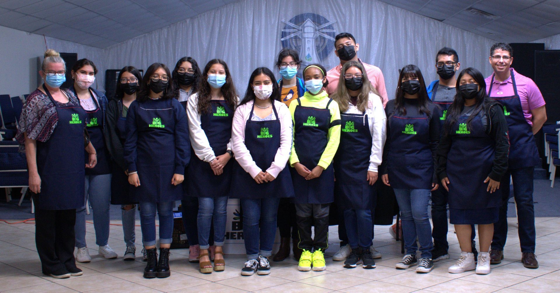 Group of people in aprons pose in front of a light-colored backdrop, likely in a room.