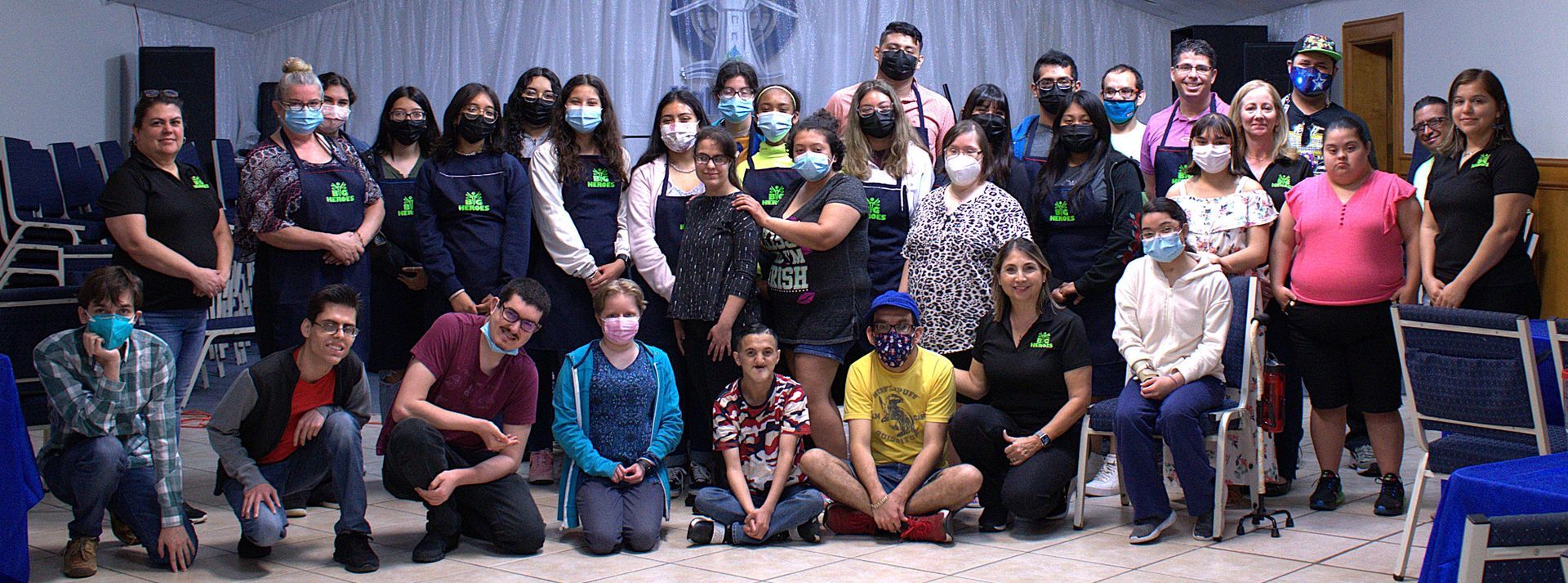 Group of people, many wearing masks, posing for a photo indoors near blue chairs.