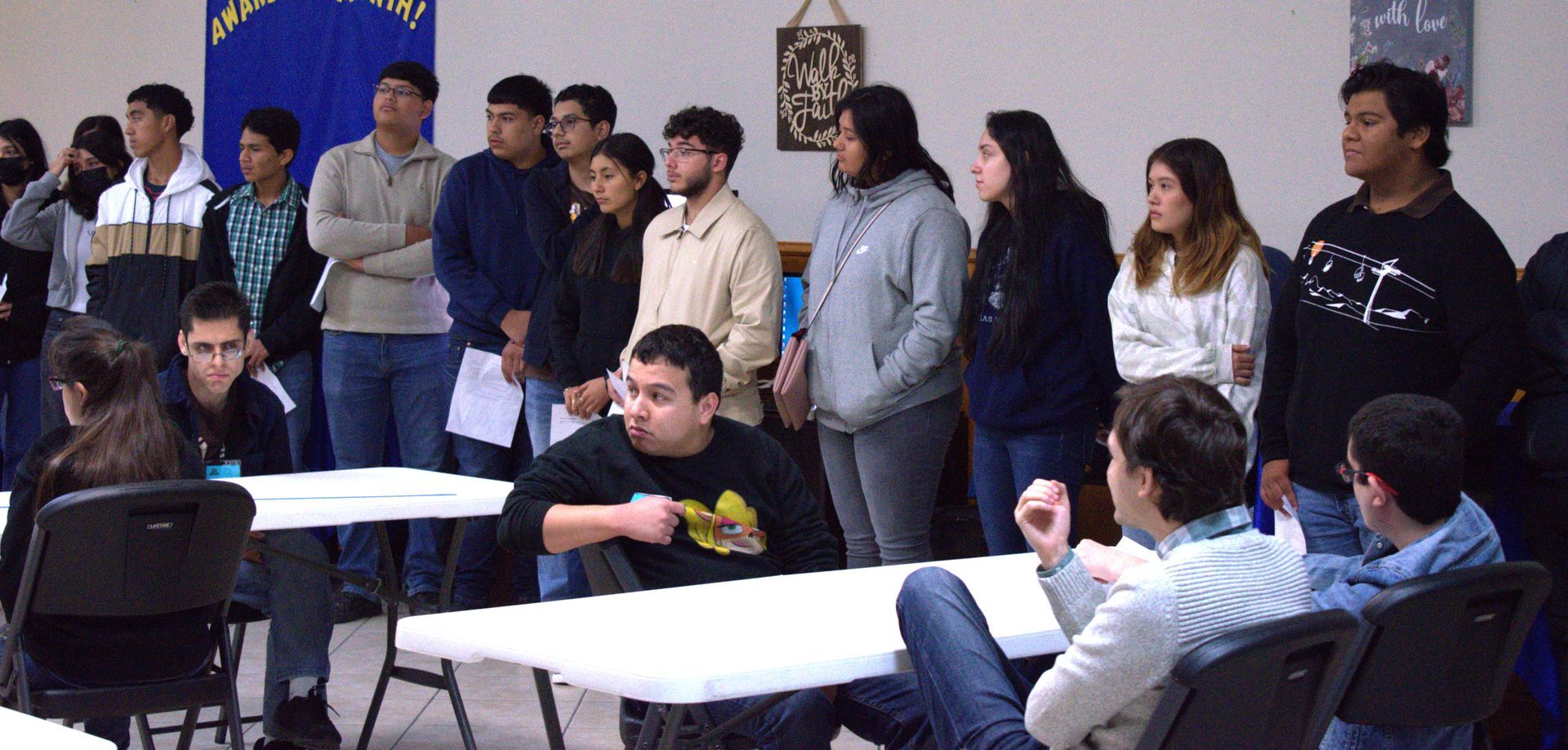 Group of students at a meeting, some standing, some seated at tables. They are inside a room.
