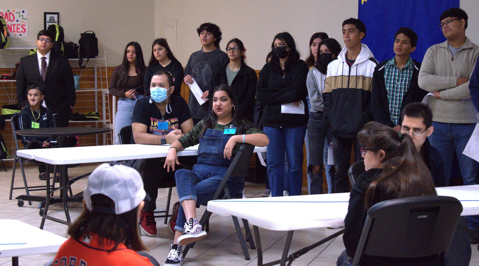 A group of diverse teens at tables, some standing, with papers. One speaks, others look on.
