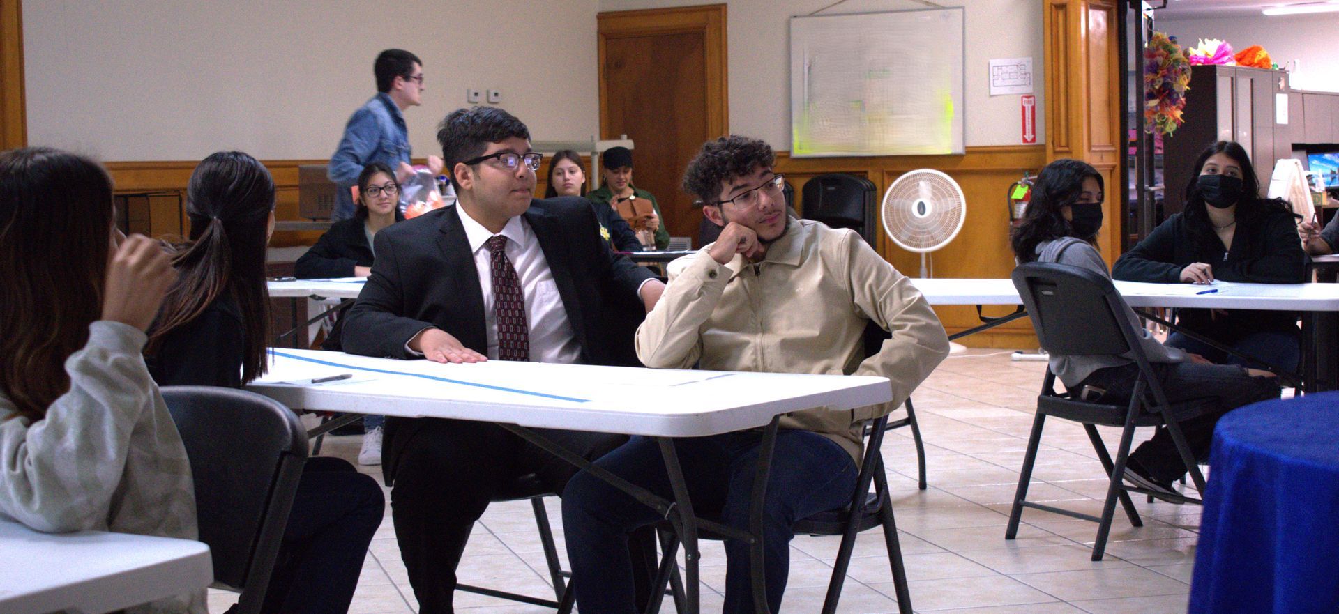 A group of people sit at tables in a room. One man in a suit and tie sits next to a man in a sweater.