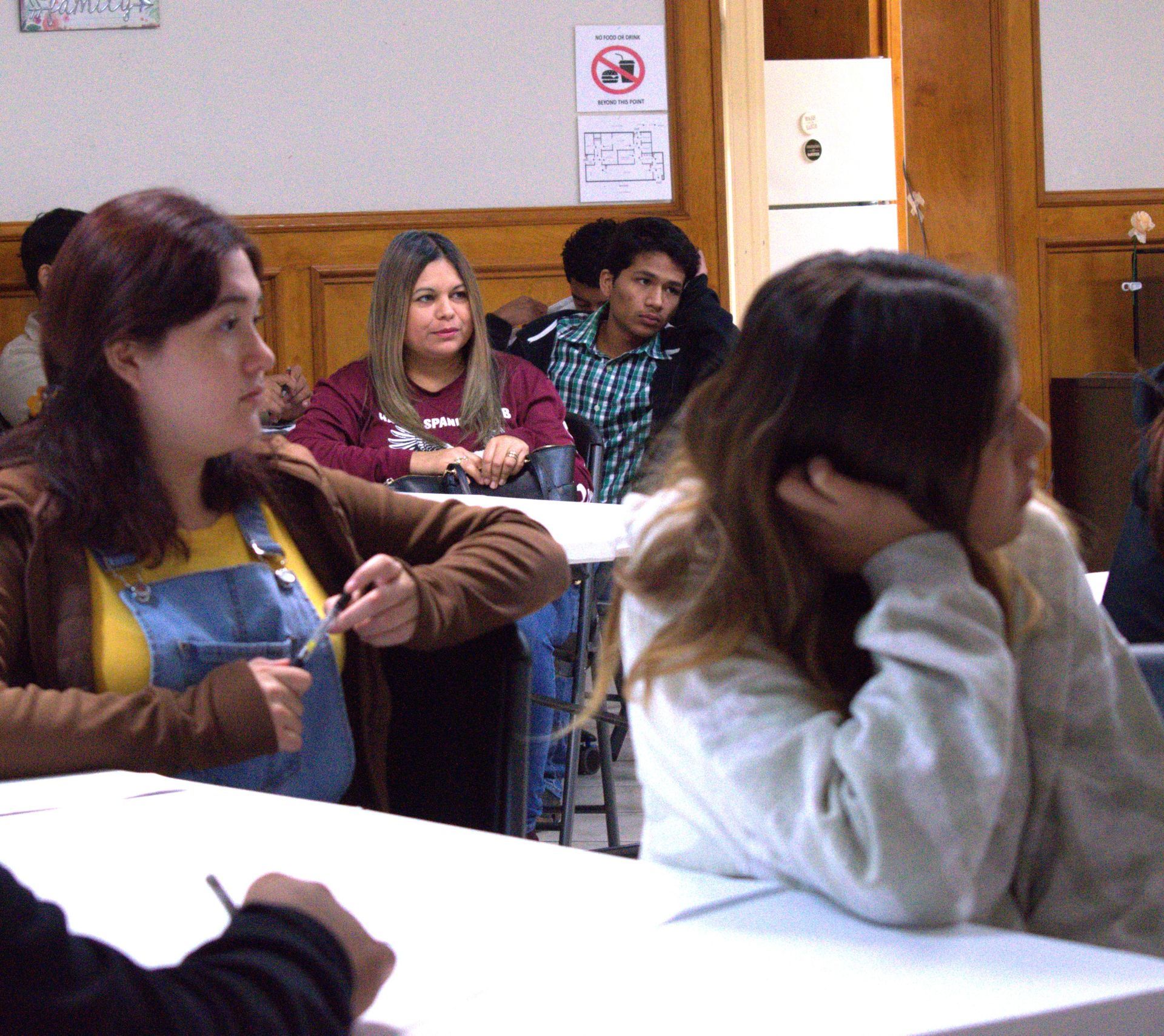Students seated in a classroom, listening. One woman points. Others look on or rest their heads.