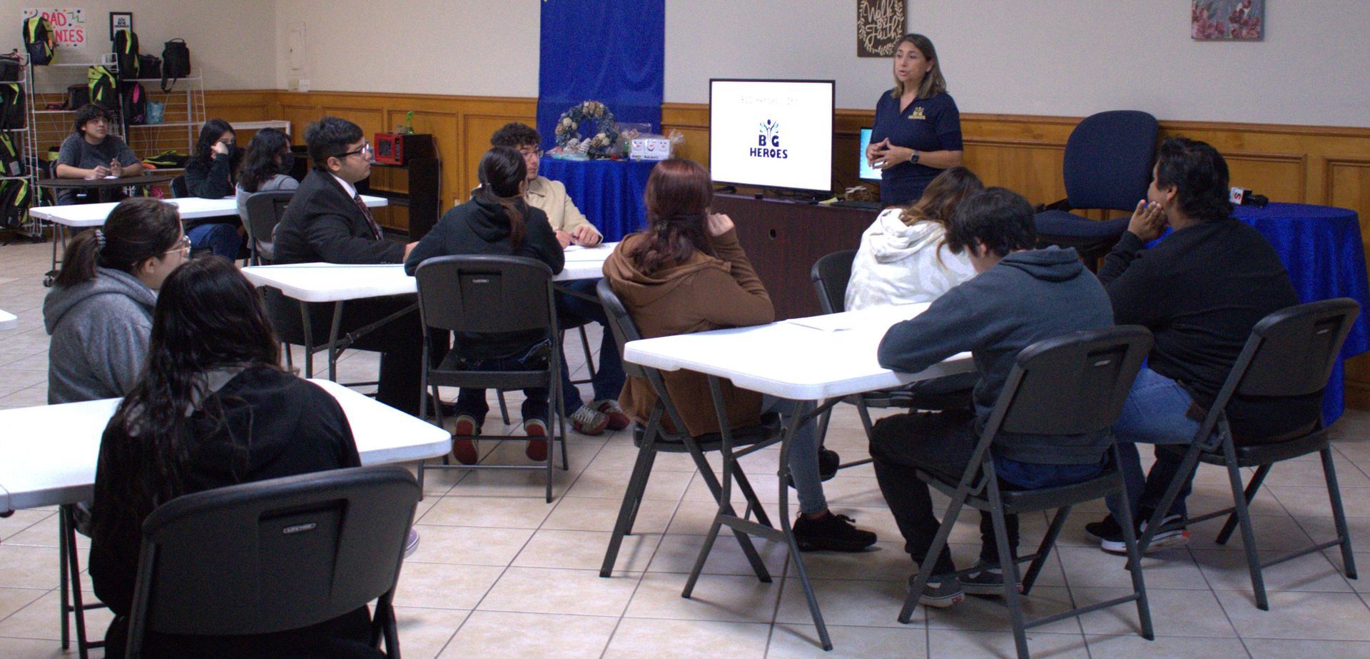A woman giving a presentation to a group of students sitting at tables in a room.