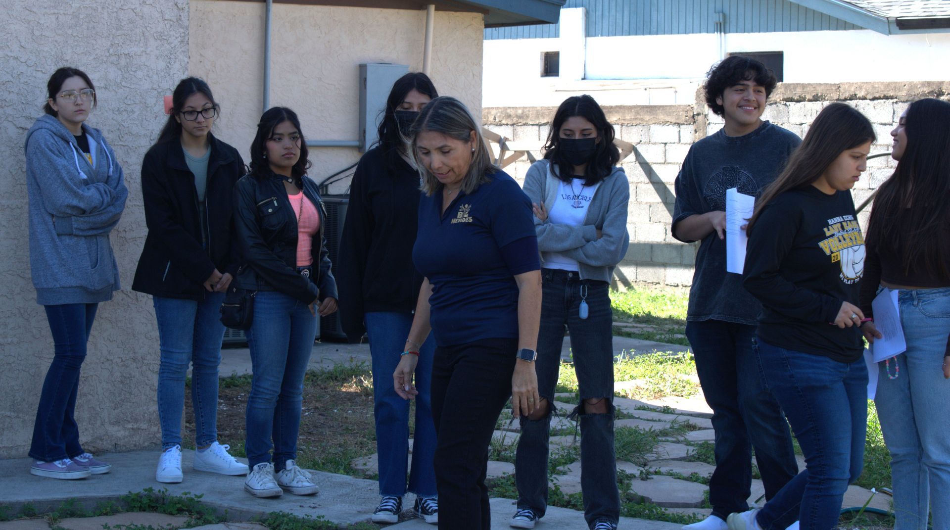 A group of students and a teacher outdoors, some wearing masks, with papers.