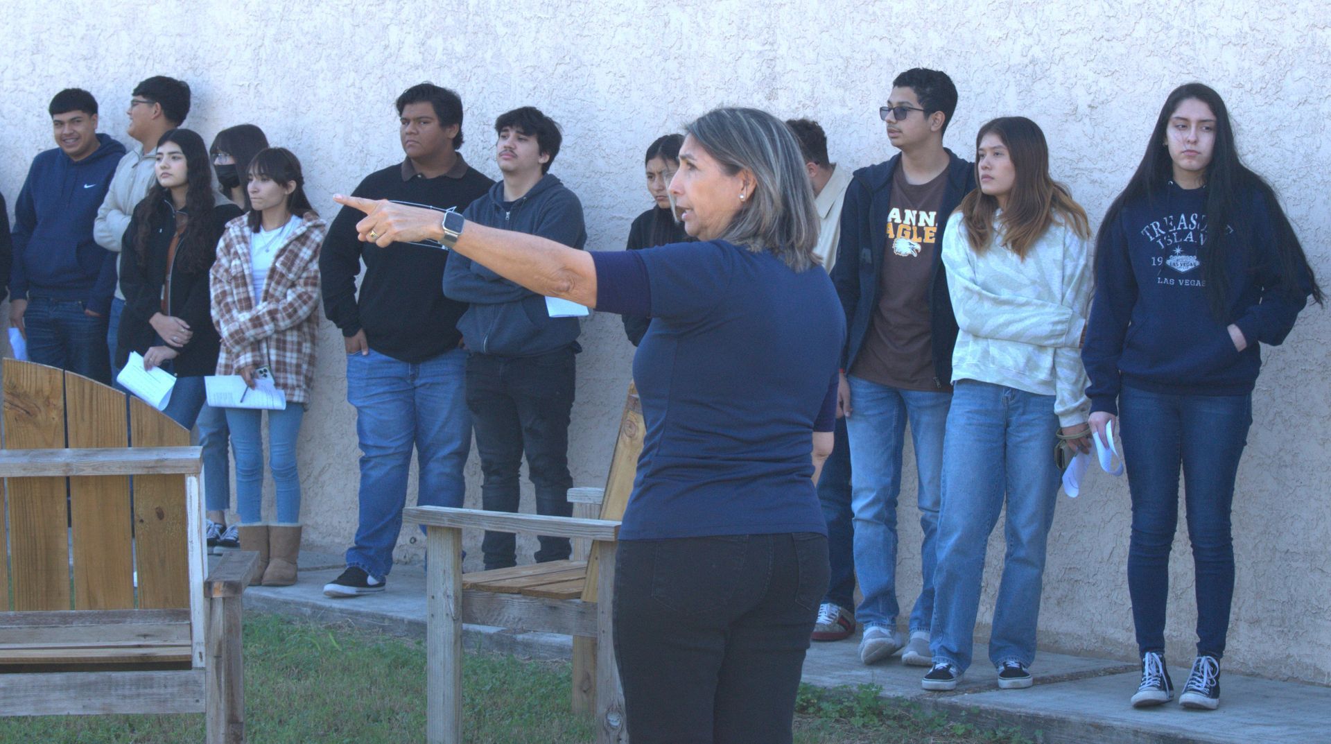 A woman gestures with her arm as she addresses a group of students outdoors near a building.