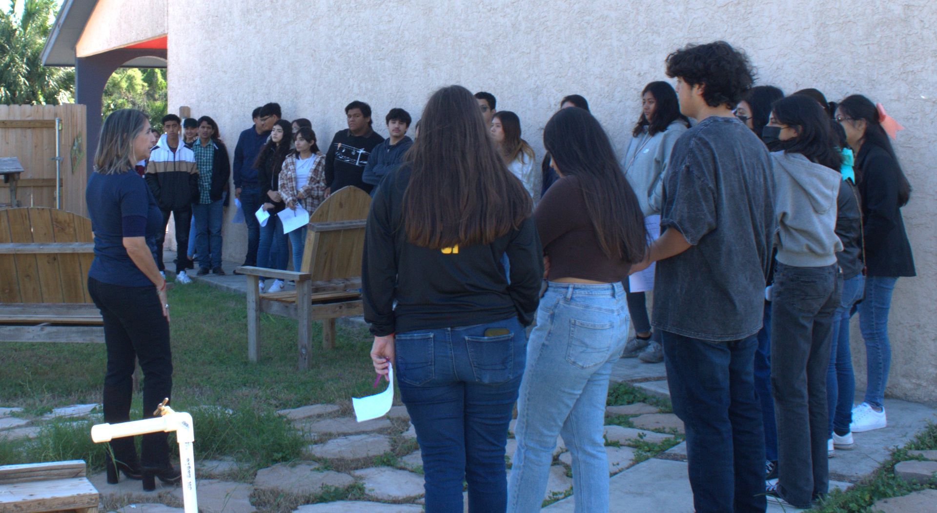 A teacher speaking to a group of students outside a building; they are listening, holding papers.