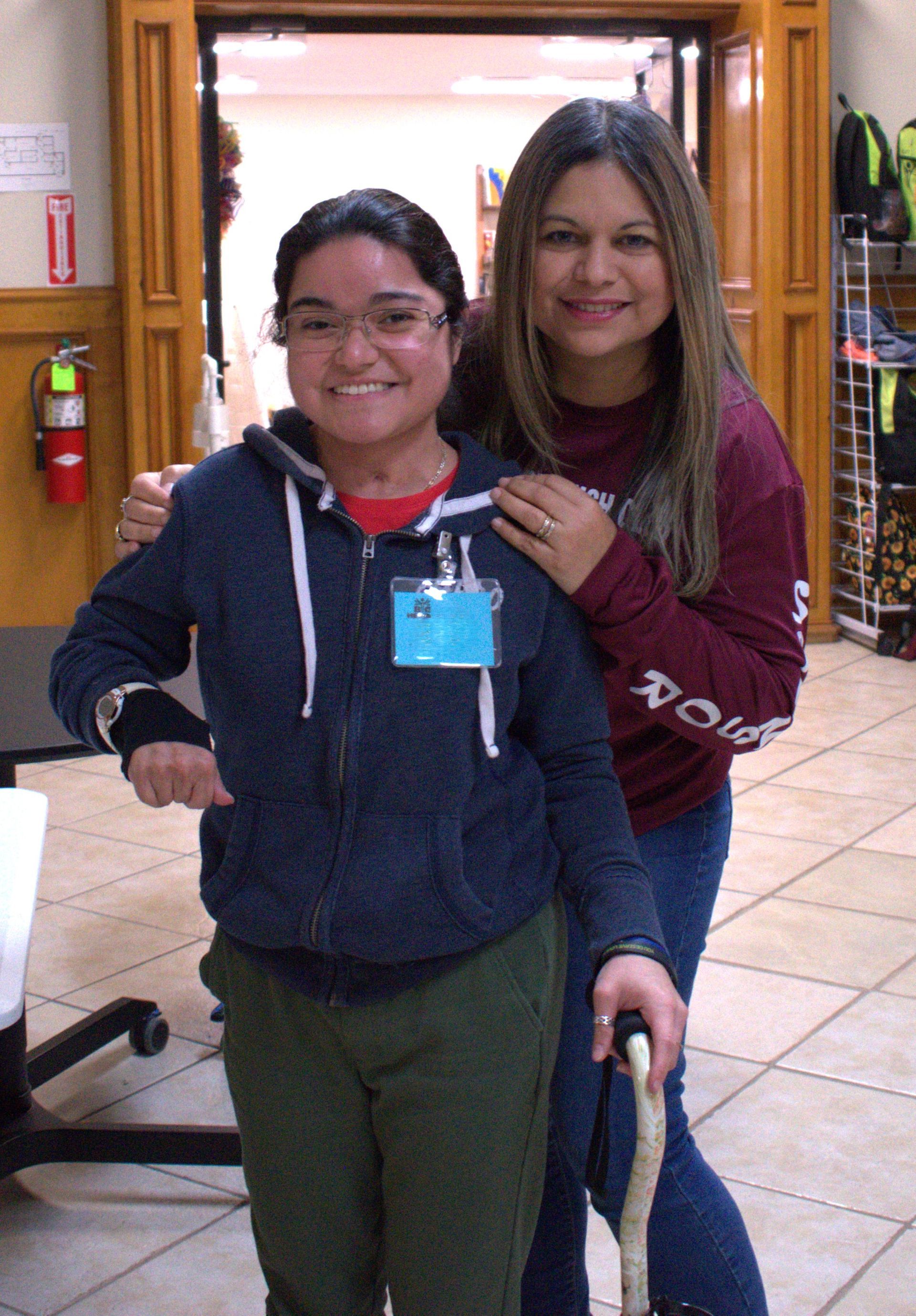 Two women posing indoors; one smiles and holds a cane, the other has an arm around her.