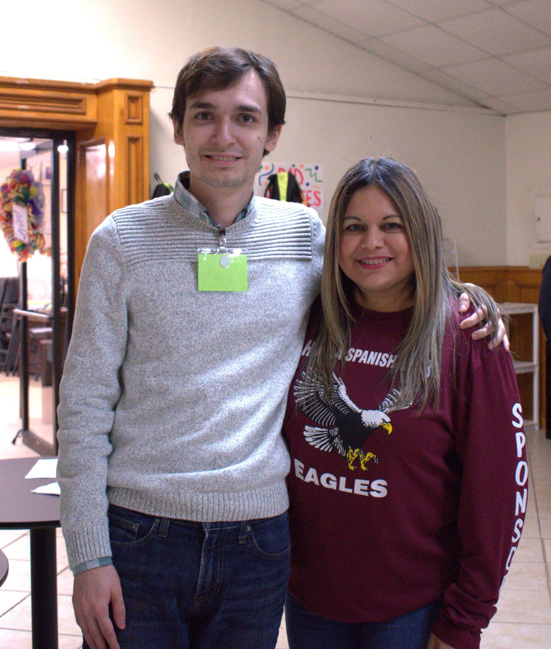 Two people, a man and woman, posing together indoors. Man in sweater, woman in maroon shirt with an eagle.