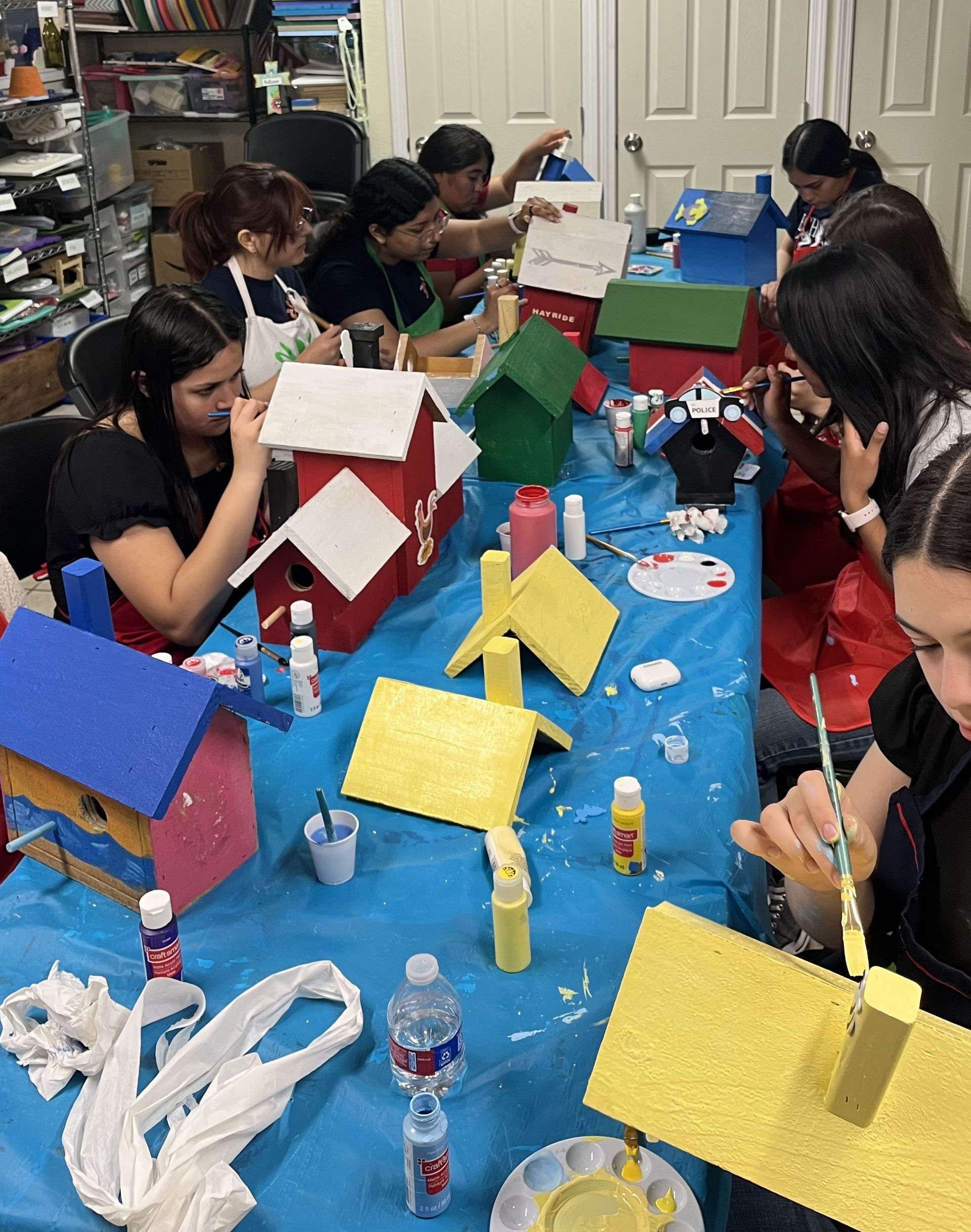 People painting birdhouses at a table. Colorful birdhouses, paint, and supplies are spread out.