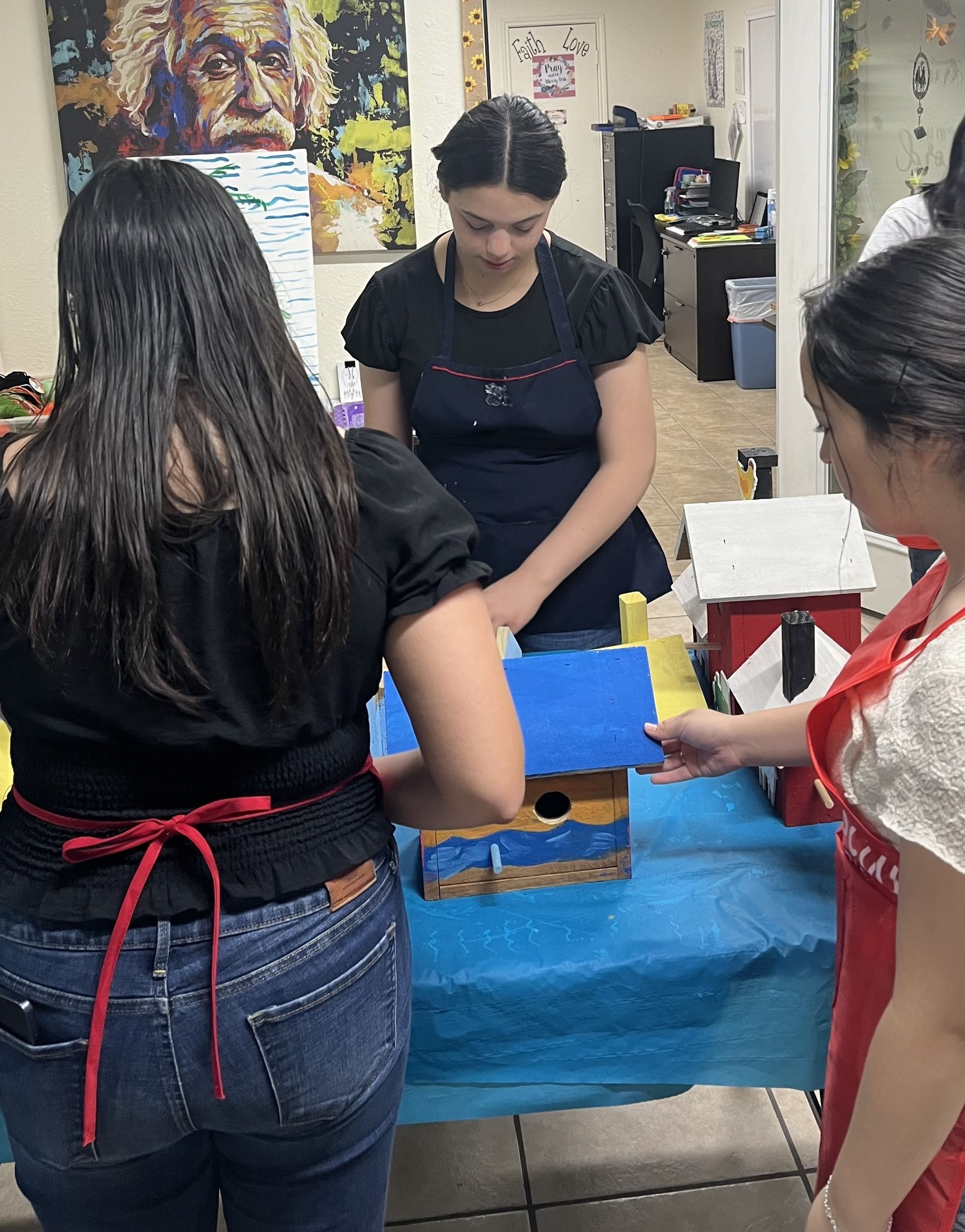 Three people paint birdhouses at a table. One wears a red apron, the other a black shirt and jeans. Indoors.
