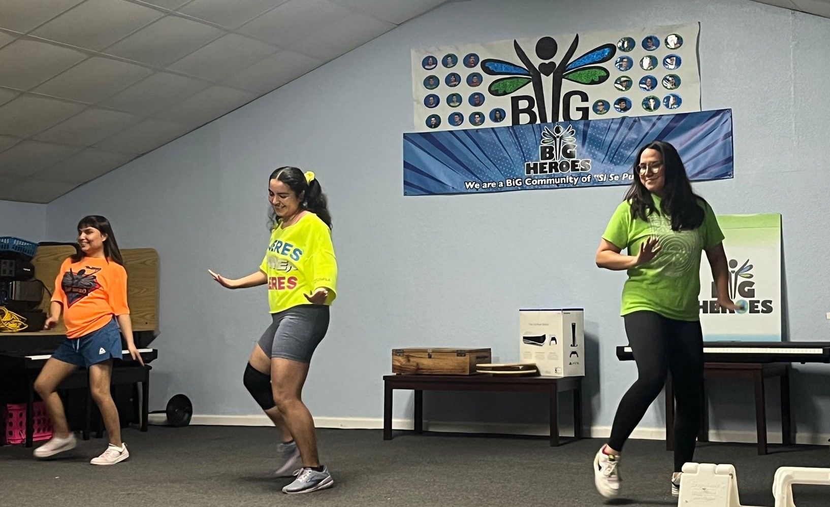 Three women are dancing in front of a sign that says big on the wall.