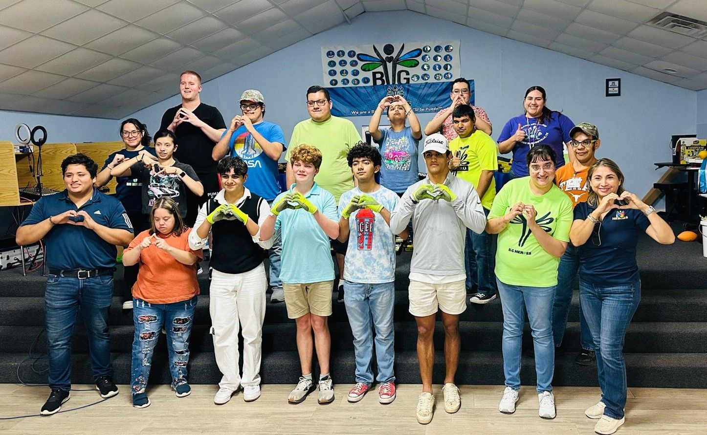 Group of people making heart shapes with their hands, in a room, under a banner.