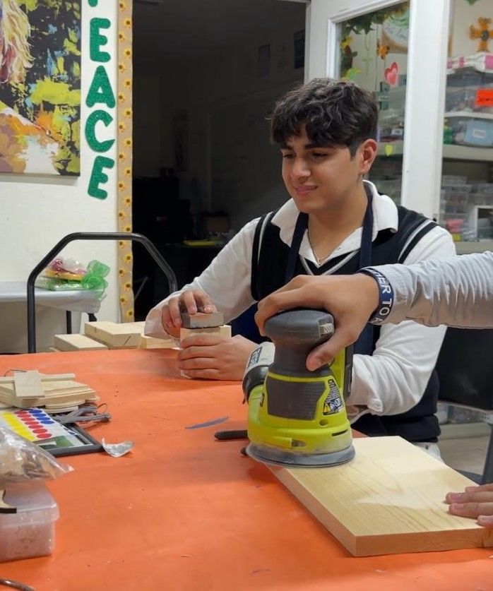 A teen sands a wooden board, smiling in a workshop with 