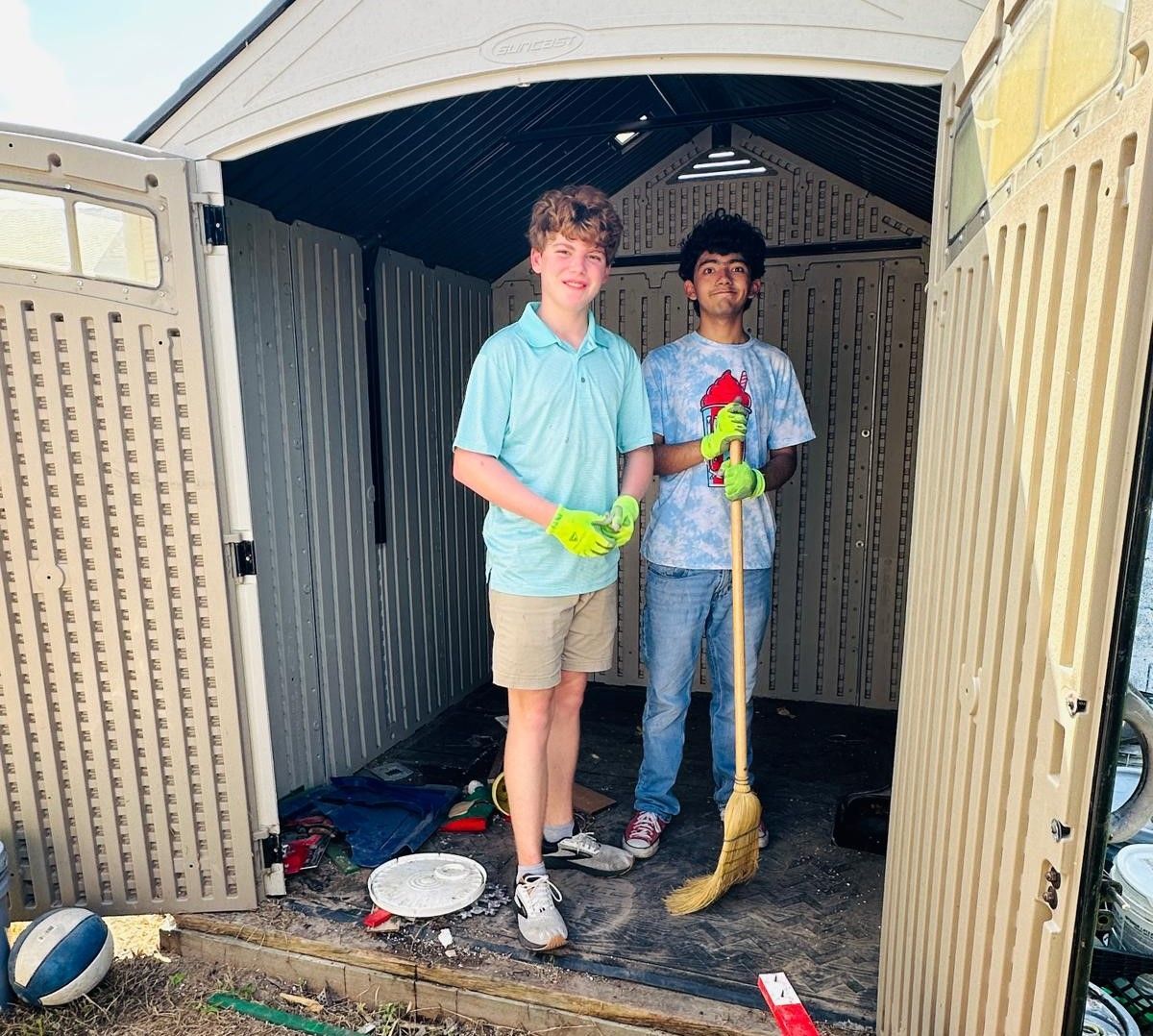 Two young men cleaning a shed; one holds a broom, both wear gloves, and smile.