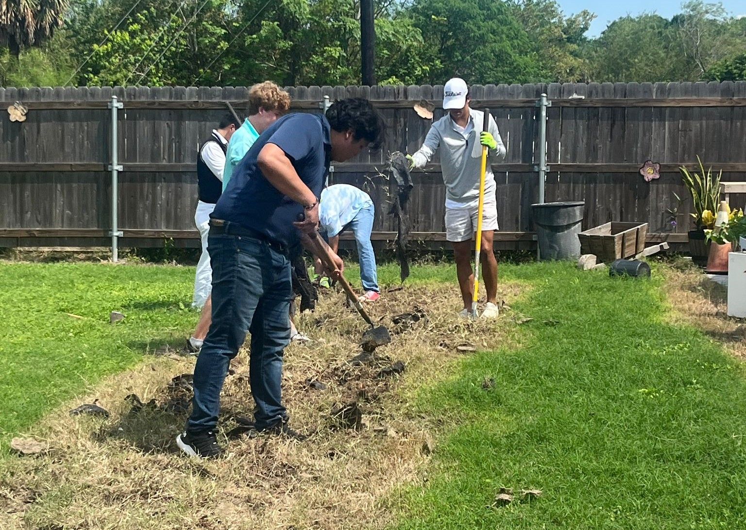 Group of people gardening in a grassy yard. They are digging in dirt under the sunlight.
