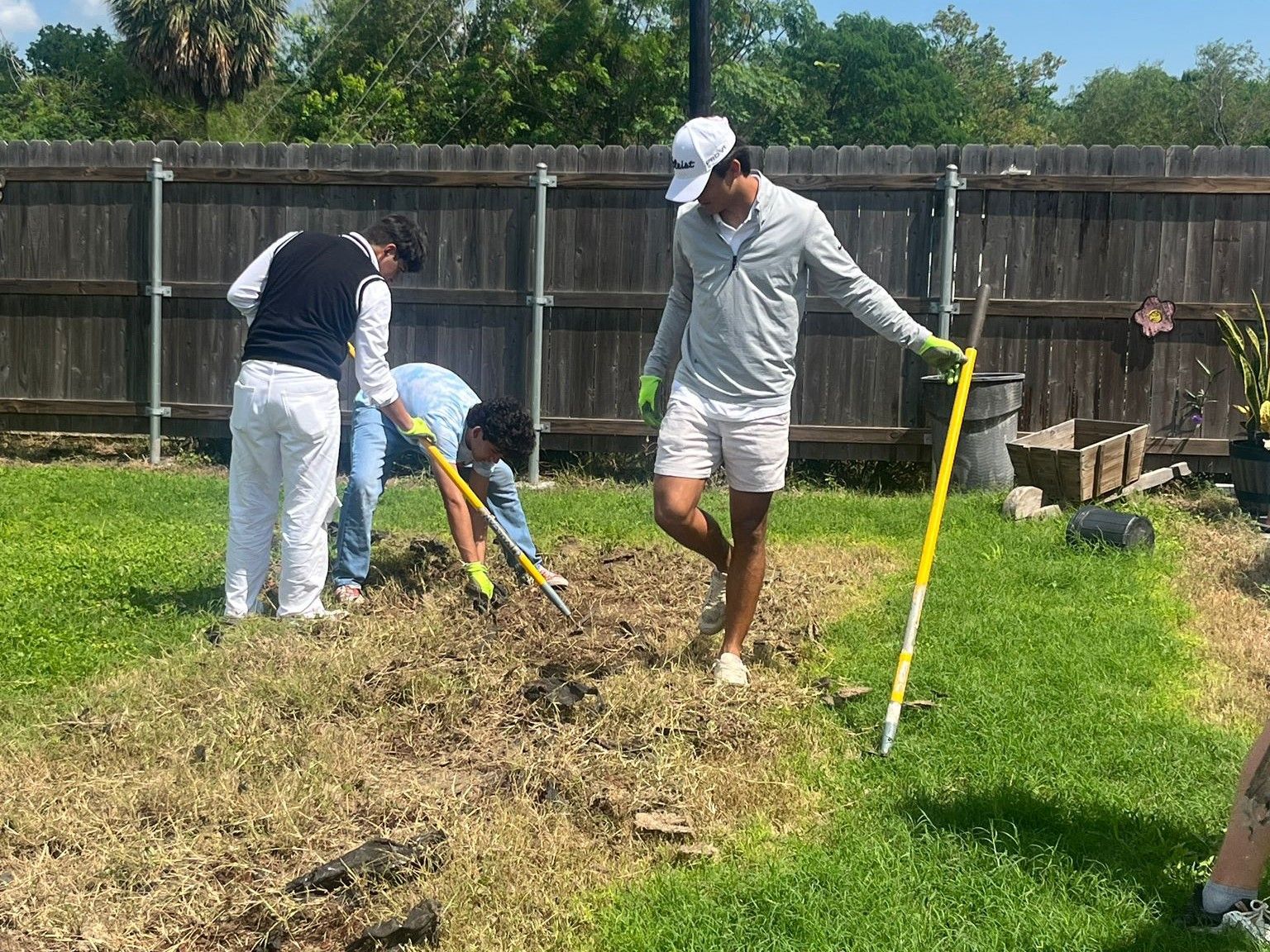 Three people gardening, removing weeds from a yard. One wears a hat and gloves, holding a tool.